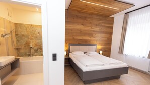 A modern bedroom with a wooden ceiling, a large bed, a nightstand, and a bathroom with a shower and sink. - Hotel Gollner (Graz)