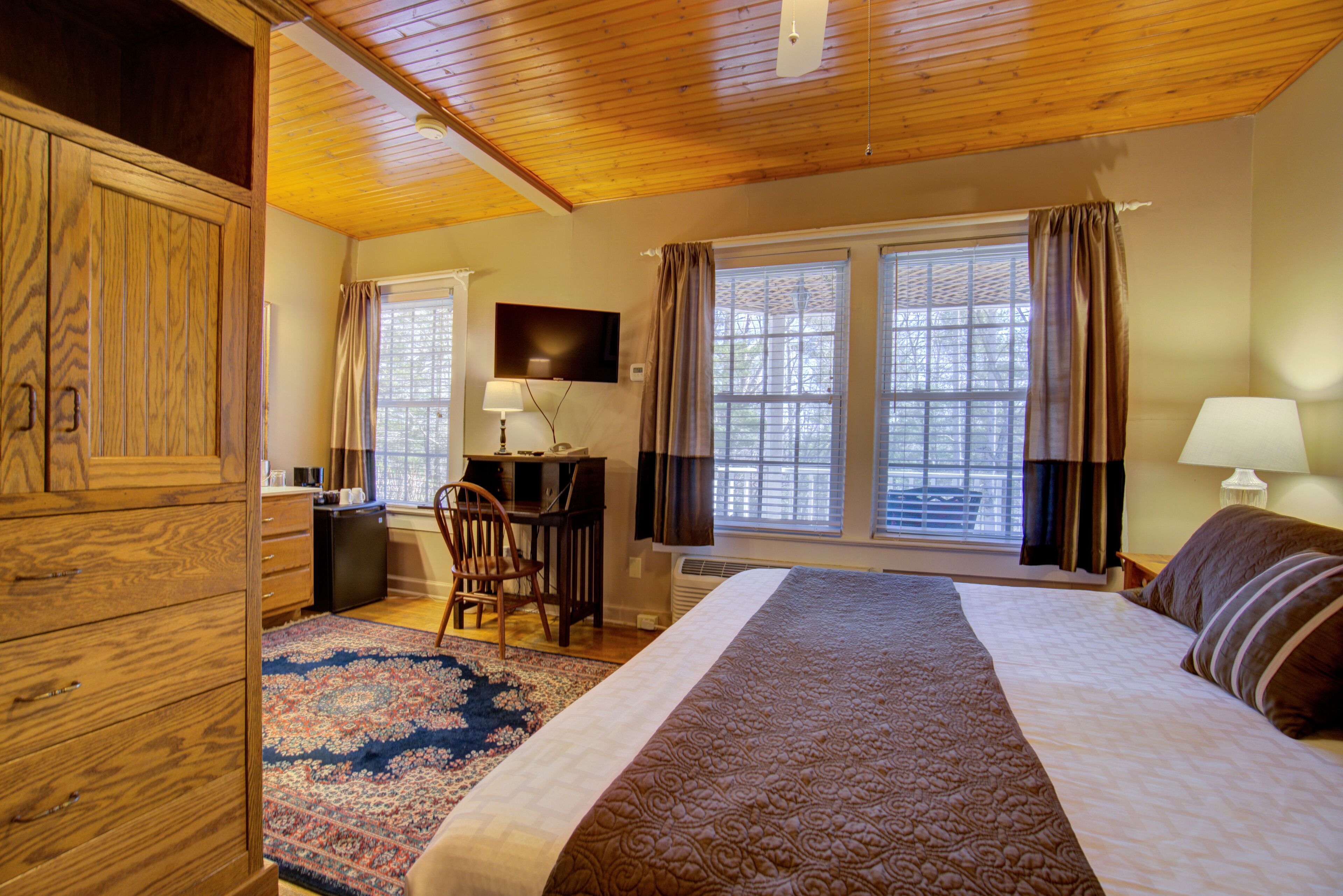 Un dormitorio con una cama grande, techo de madera, un escritorio con silla, televisor y ventana con cortinas.