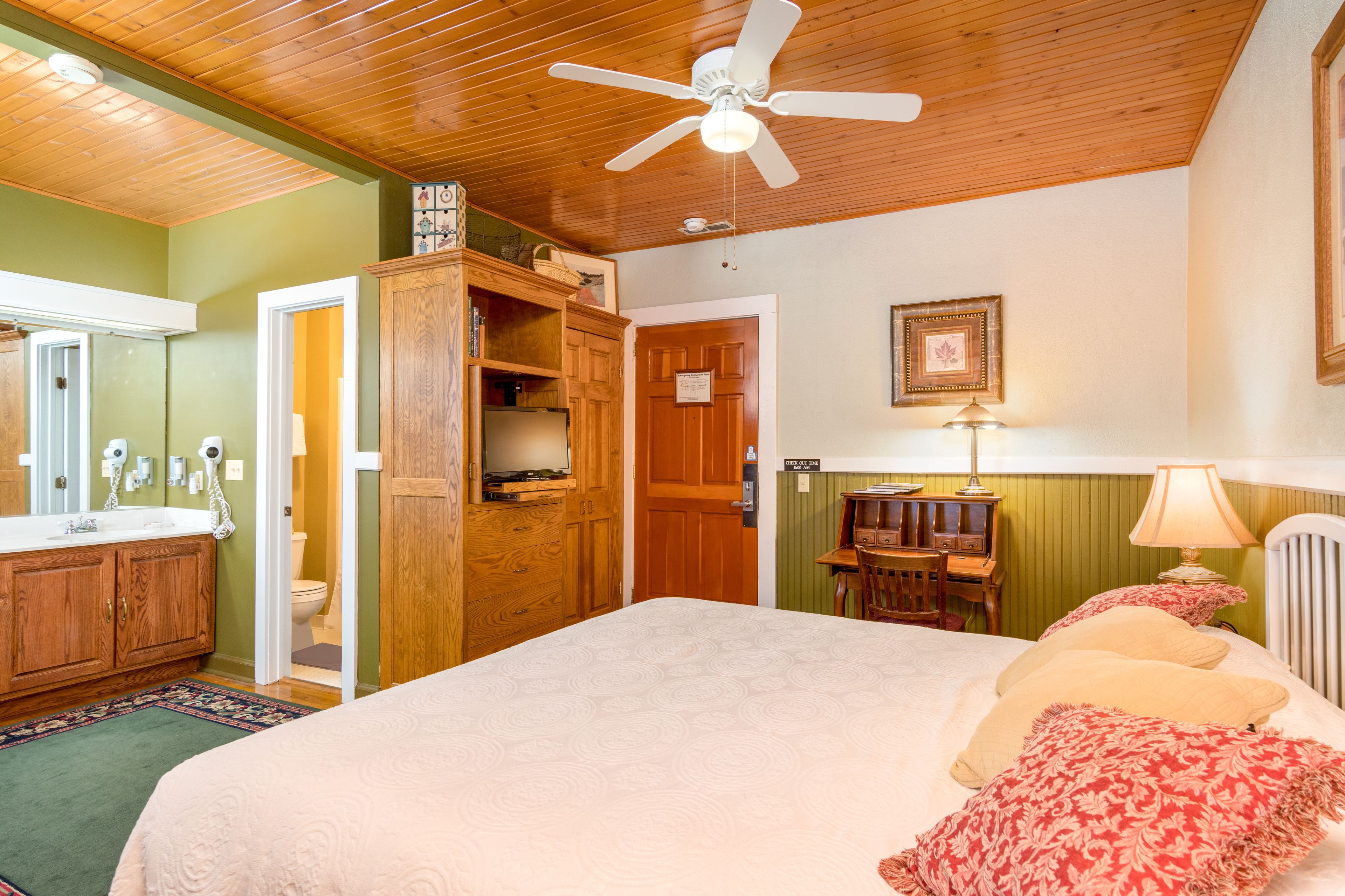 Ein Schlafzimmer mit einem großen Bett, Holzboden, einem Deckenventilator, einem Schrank, einem Schreibtisch mit Stuhl, einer Lampe und einem gerahmten Bild an der Wand.
