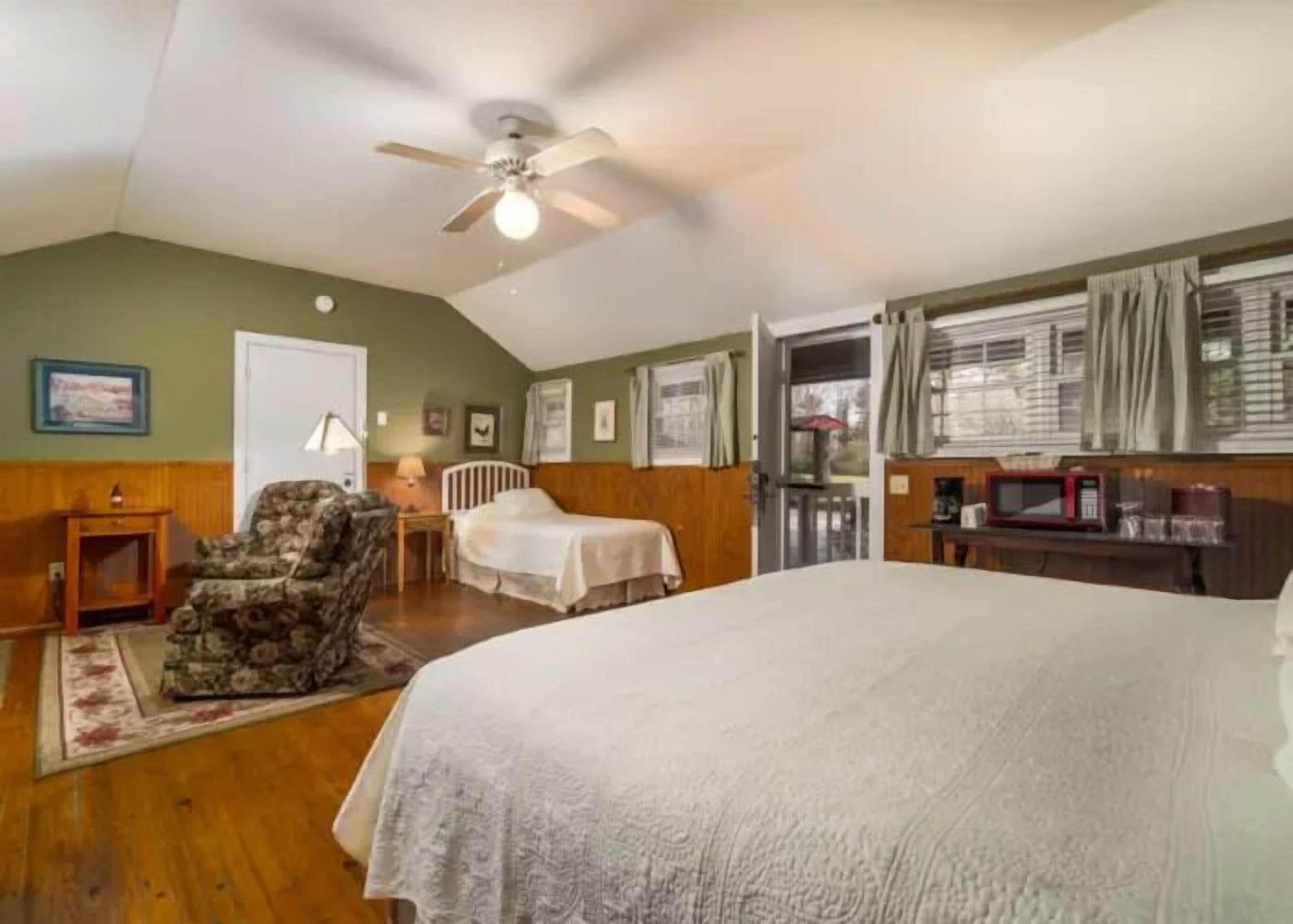 A bedroom with a large bed, a chair, a desk, a ceiling fan, and a door leading to a balcony.