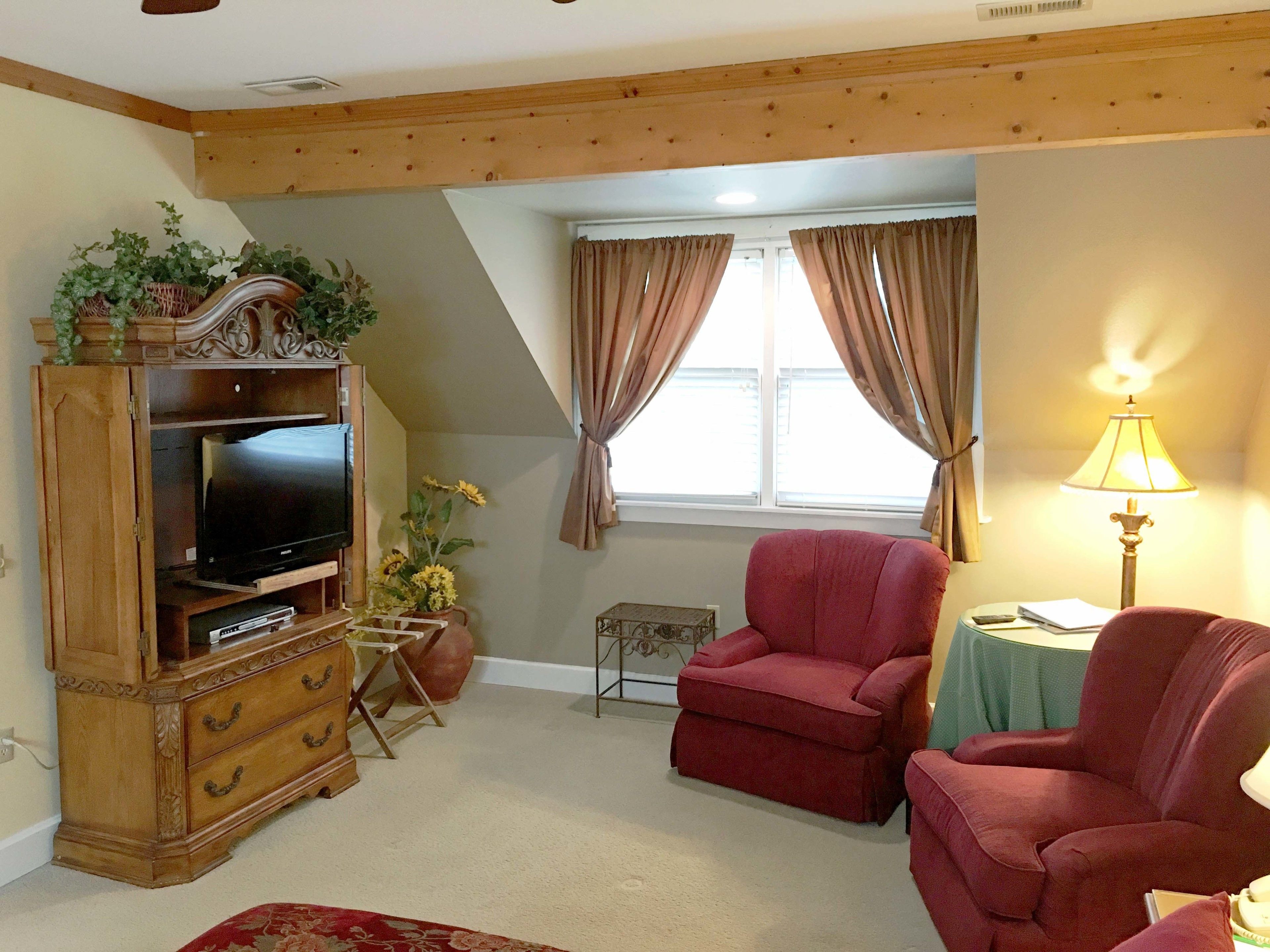 Ein gemütliches Wohnzimmer mit Fernseher auf einer Holzkommode, einem roten Sessel, einem kleinen Tisch mit Lampe und einem Fenster mit Vorhängen.