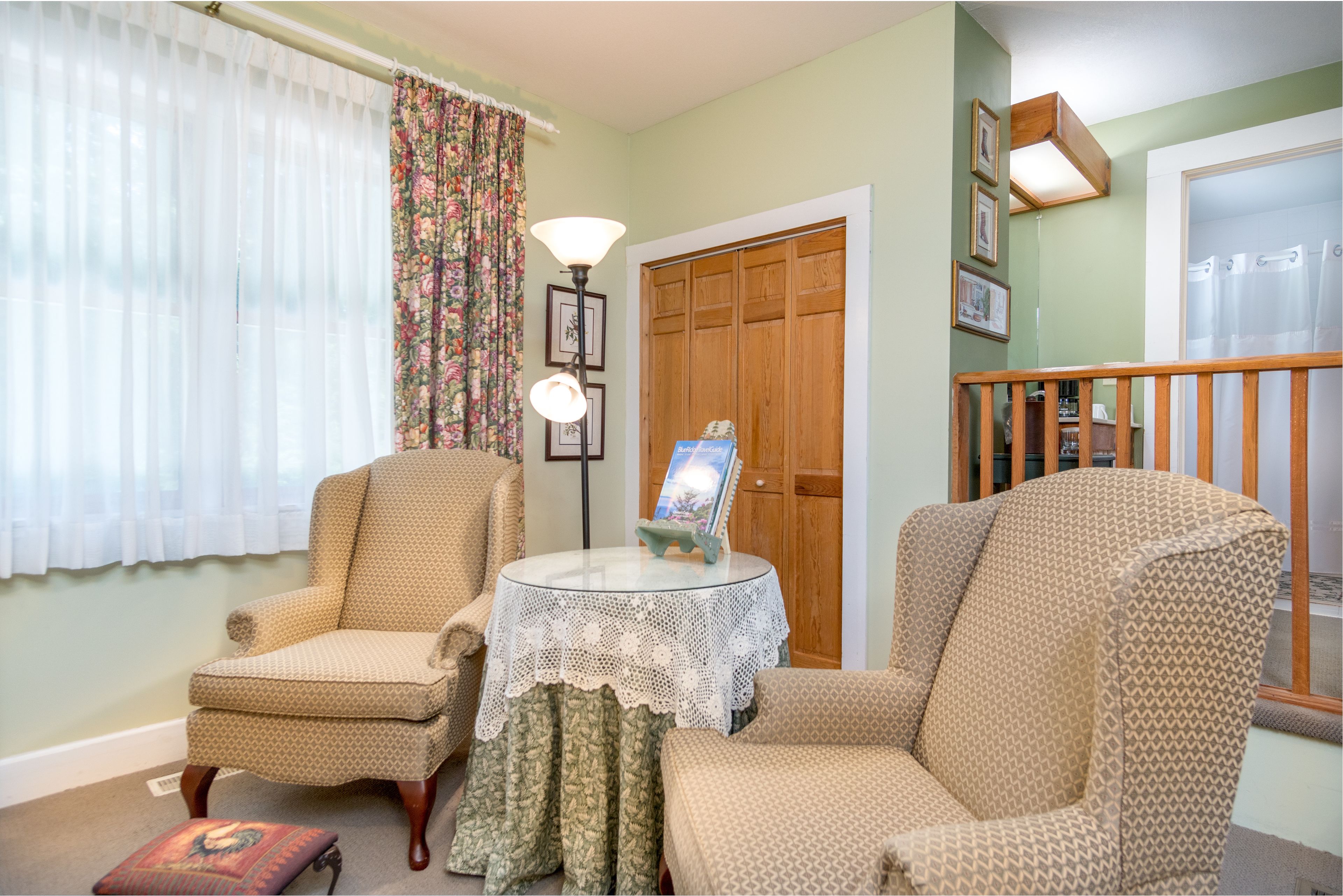 A cozy living room with two patterned armchairs, a small round table with a lace tablecloth, and a wooden door in the background.