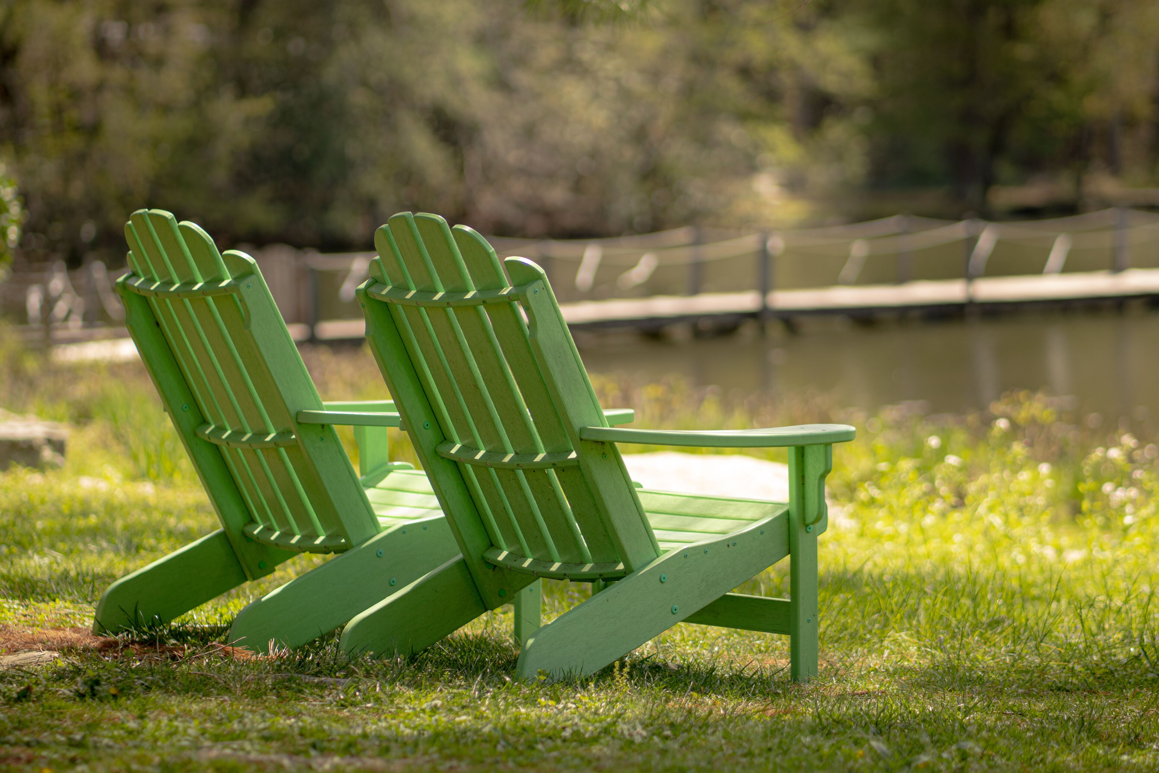 Two green Adirondack chairs positioned on grass near a body of water with a wooden bridge in the background.