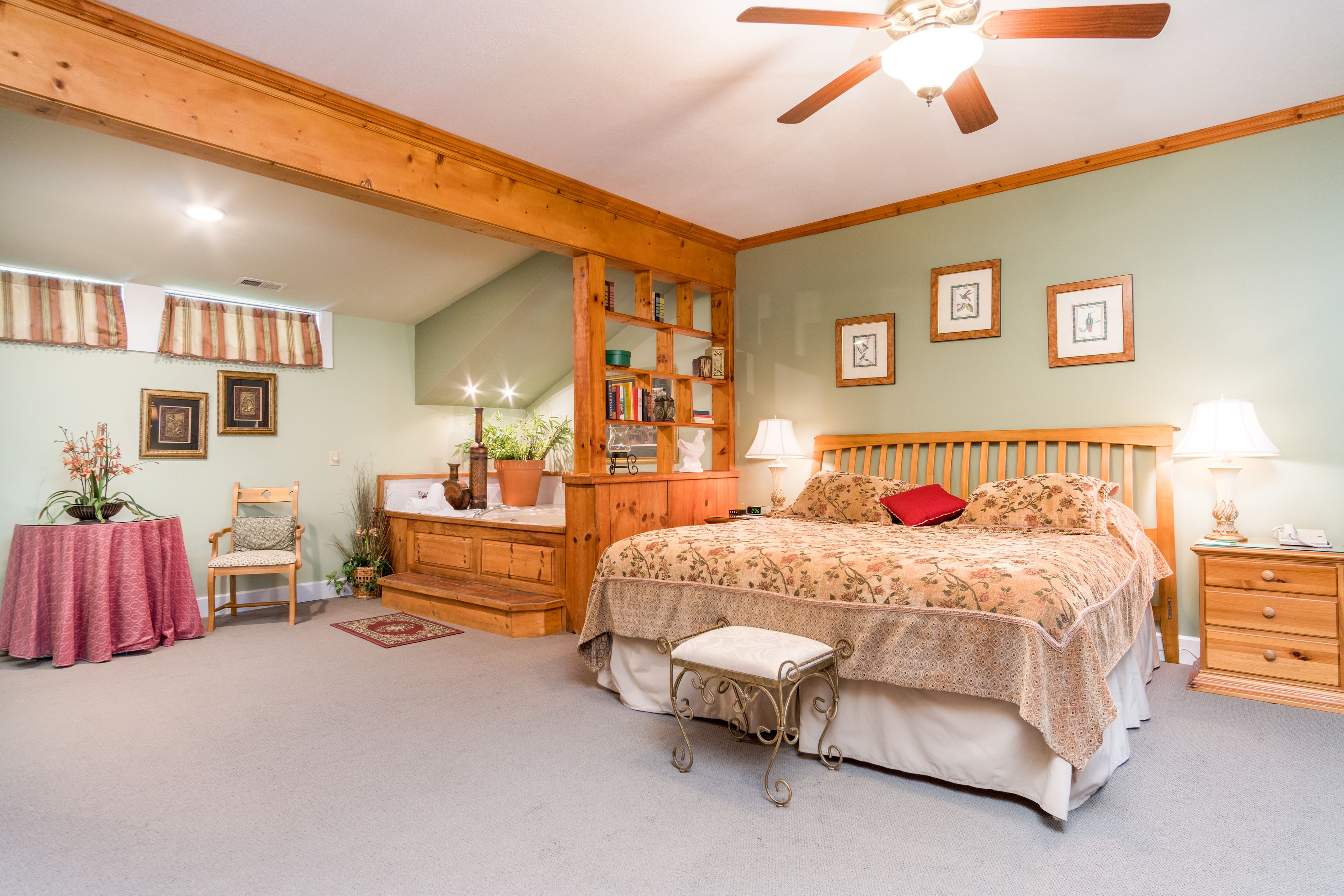 A bedroom with a bed, bedside tables, a wooden headboard, a ceiling fan, a bookshelf, and a small table with a chair.