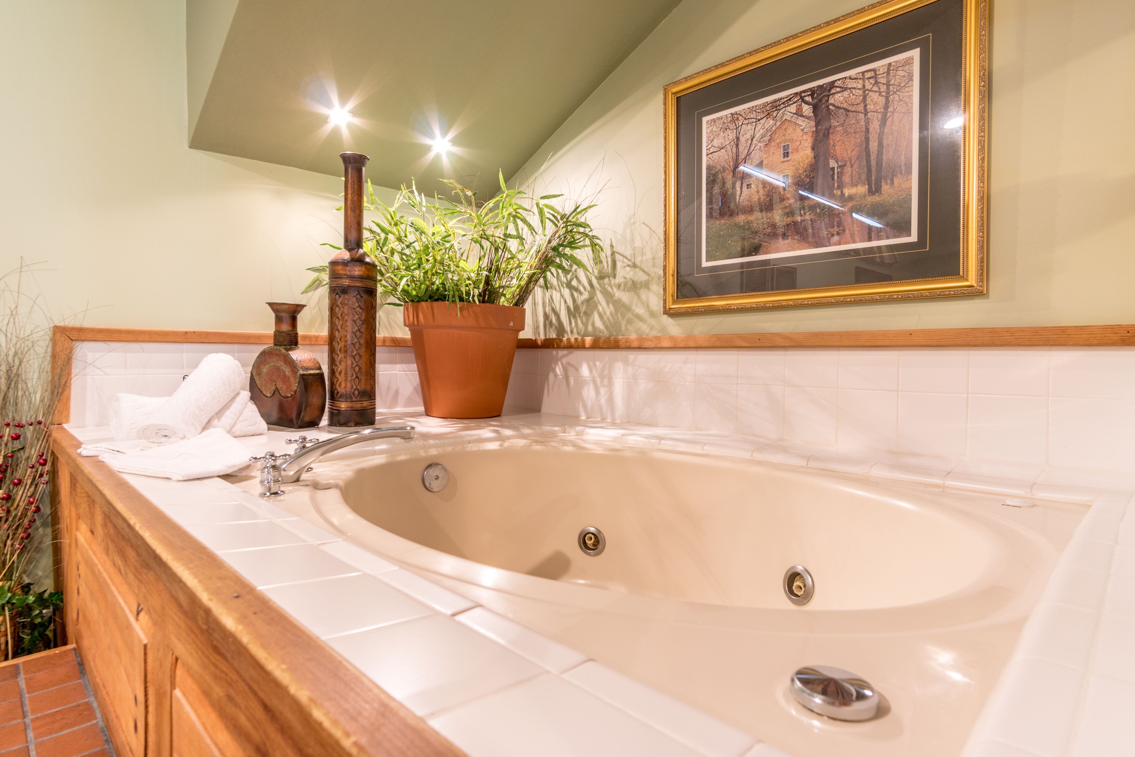 A bathroom with a large bathtub, a wooden vanity, a potted plant, and a framed picture on the wall.