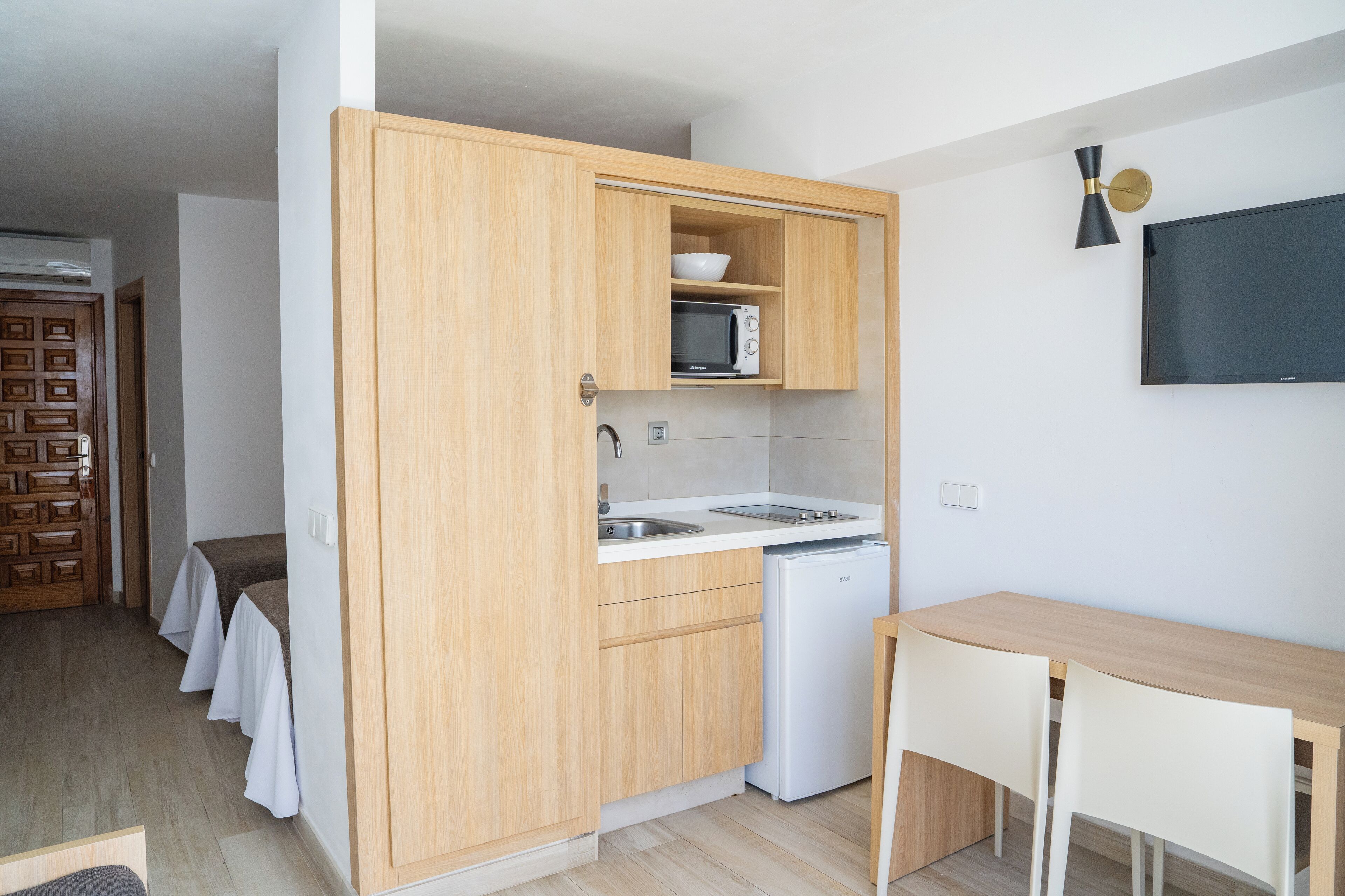 A compact kitchen with wooden cabinets, a microwave, a sink, and a small refrigerator. There is a wooden table and chairs in the adjacent dining area.