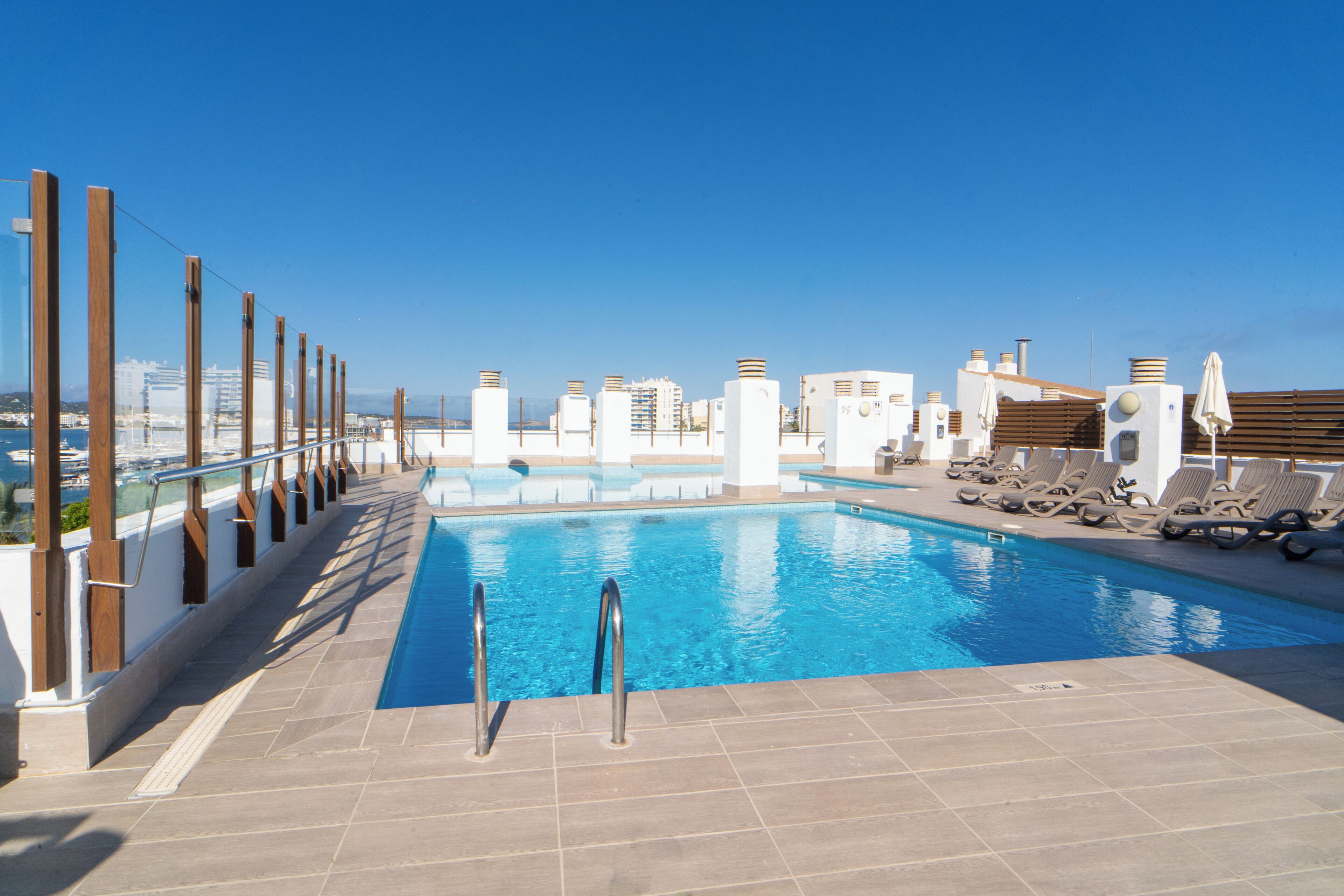 Una piscina en la azotea con cielo azul despejado, terraza de baldosas y sillas de descanso.