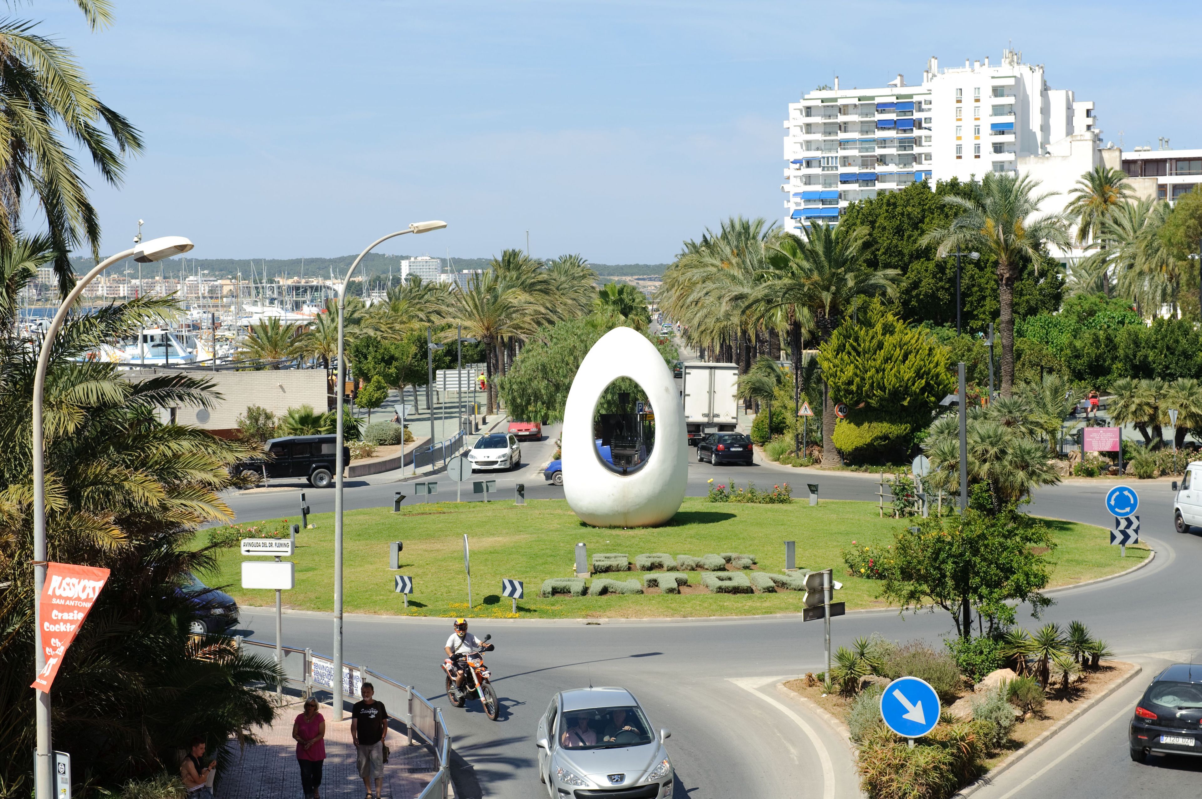 A roundabout with a distinctive white sculpture, palm trees, and a modern building in the background.
