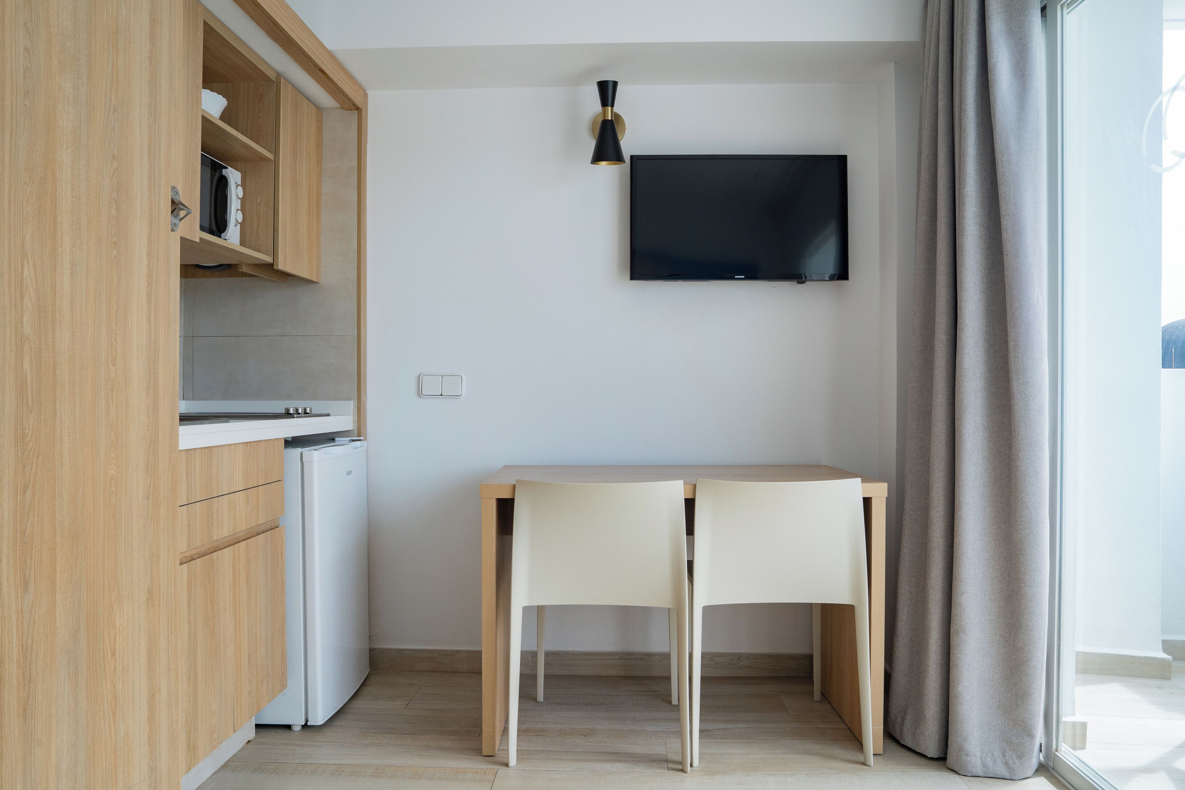 A compact kitchen with a built-in microwave, a small refrigerator, and a wooden cabinet. A flat-screen TV is mounted on the wall above a wooden dining table with two chairs.