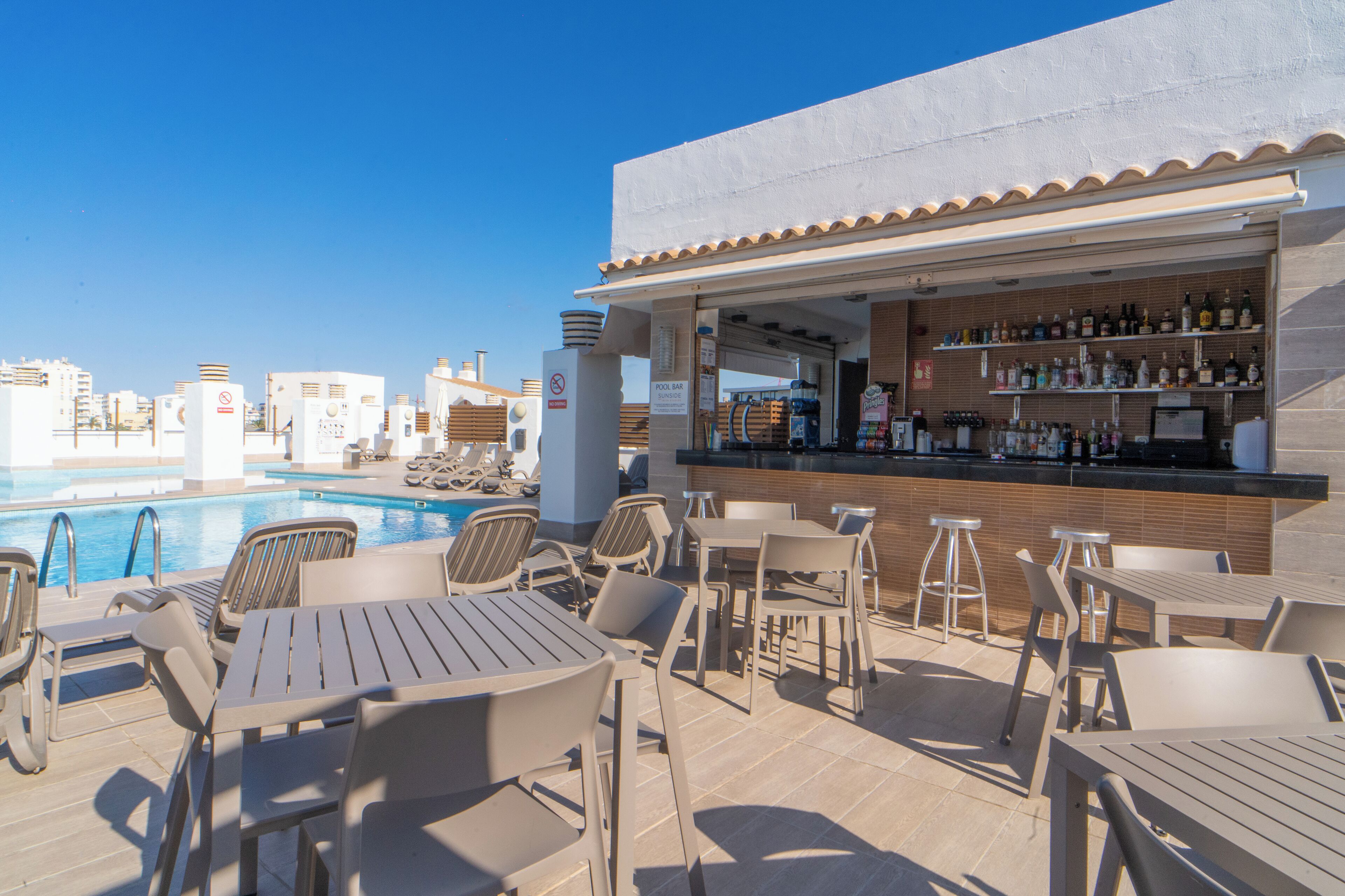 Un bar situé sur le toit, offrant une vue sur la piscine, avec des places assises en plein air et un comptoir bien garni de différentes bouteilles.