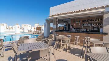 Un bar situé sur le toit, offrant une vue sur la piscine, avec des places assises en plein air et un comptoir bien garni de différentes bouteilles.