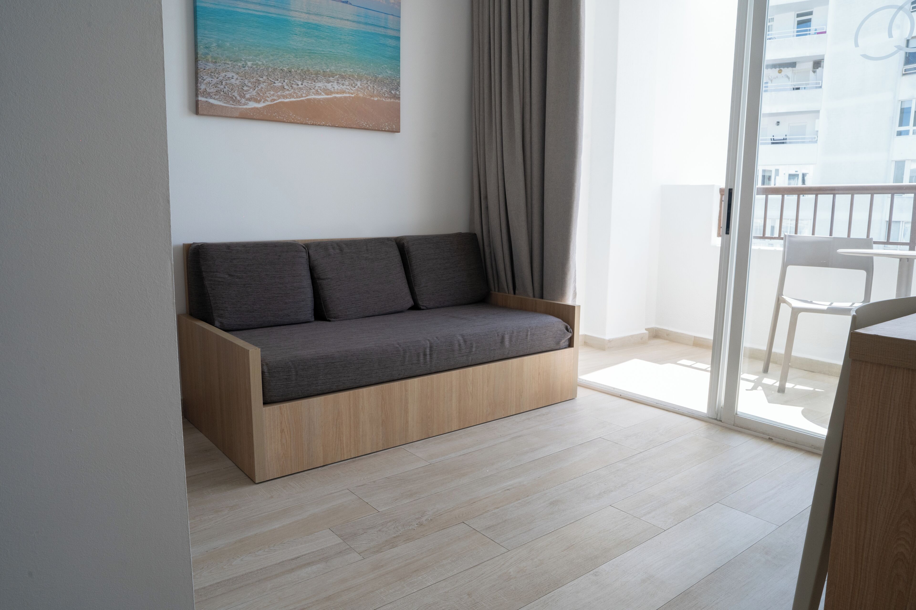 Una sala de estar moderna con un sofá de madera, un cuadro de una playa y vistas a un balcón con una silla.
