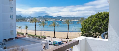 A balcony with a view of a beach, palm trees, and a distant cityscape.