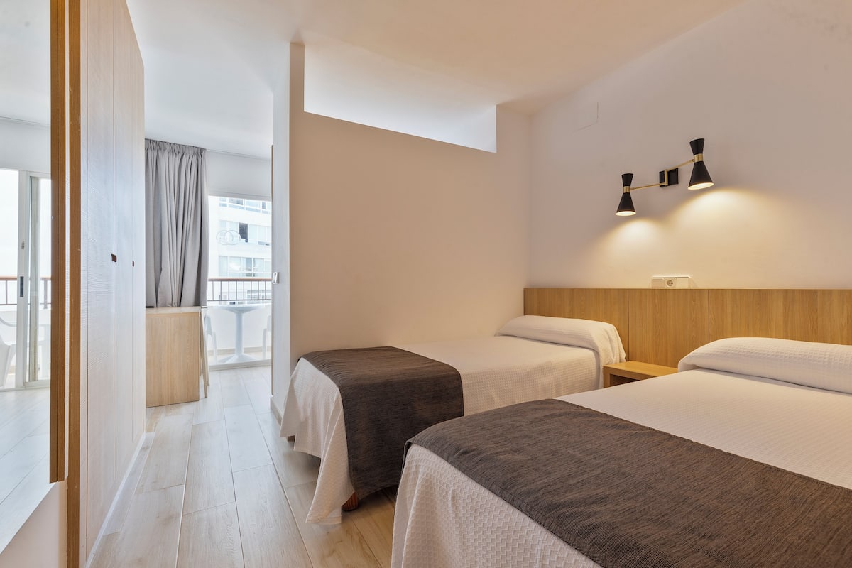 A hotel room with two beds, a wooden headboard, a nightstand, and a wall-mounted lamp.