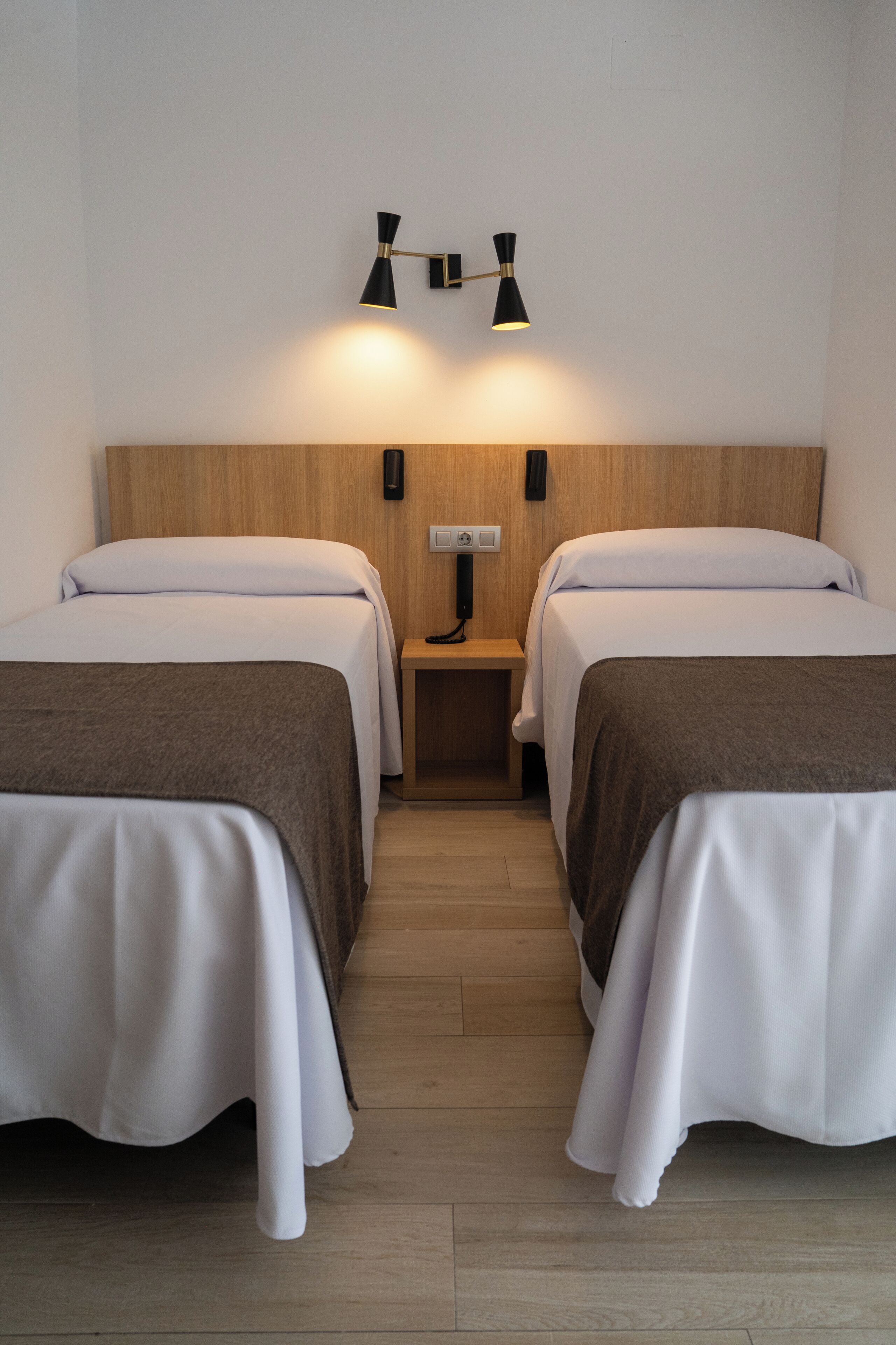 A hotel room with two beds, a wooden headboard, a nightstand with a lamp, and a wall socket.