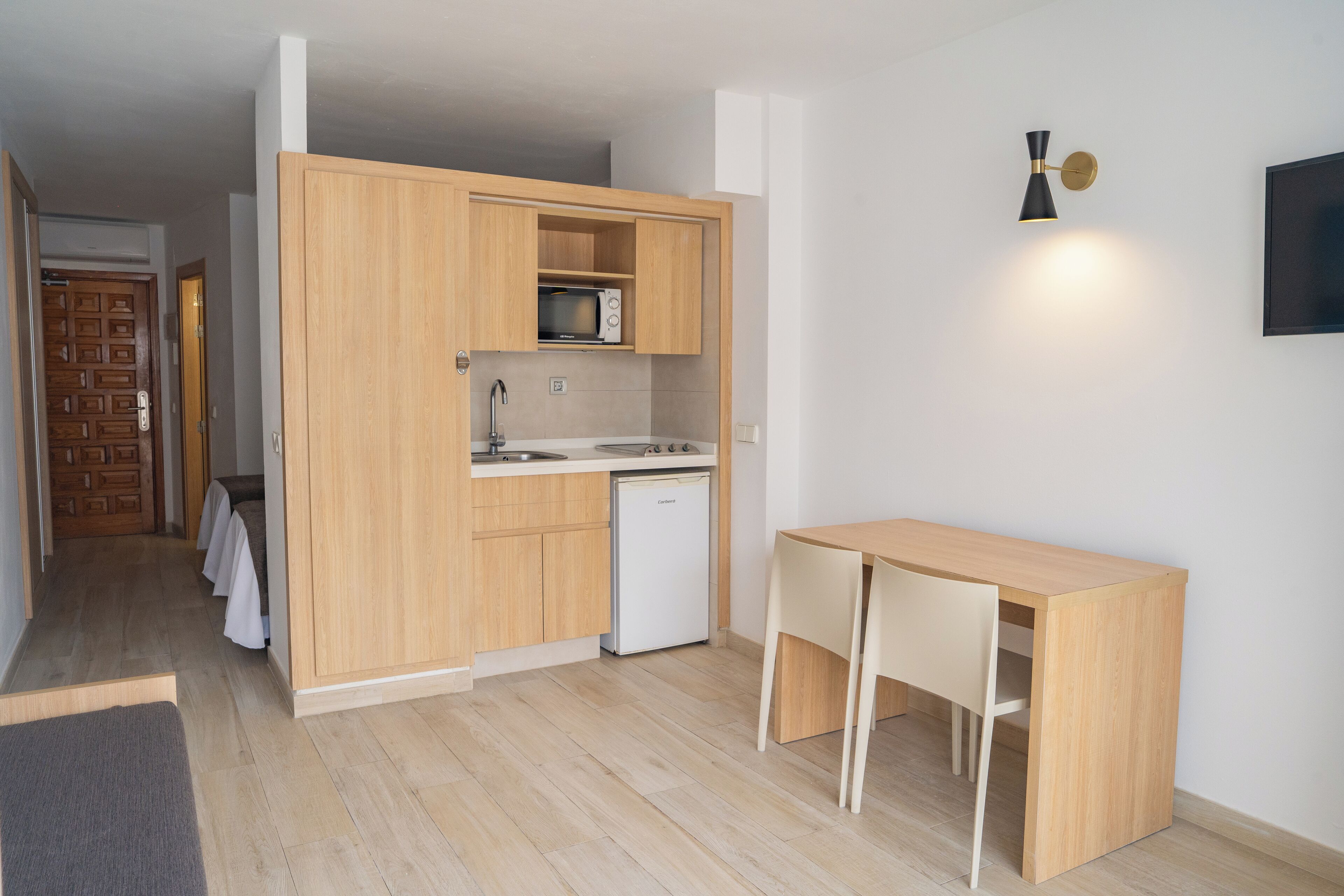 Cocina compacta con armarios de madera, microondas, lavamanos y un refrigerador pequeño. Hay una mesa de madera con sillas.
