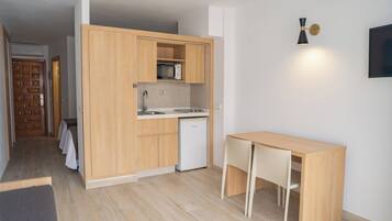 Cocina compacta con armarios de madera, microondas, lavamanos y un refrigerador pequeño. Hay una mesa de madera con sillas.