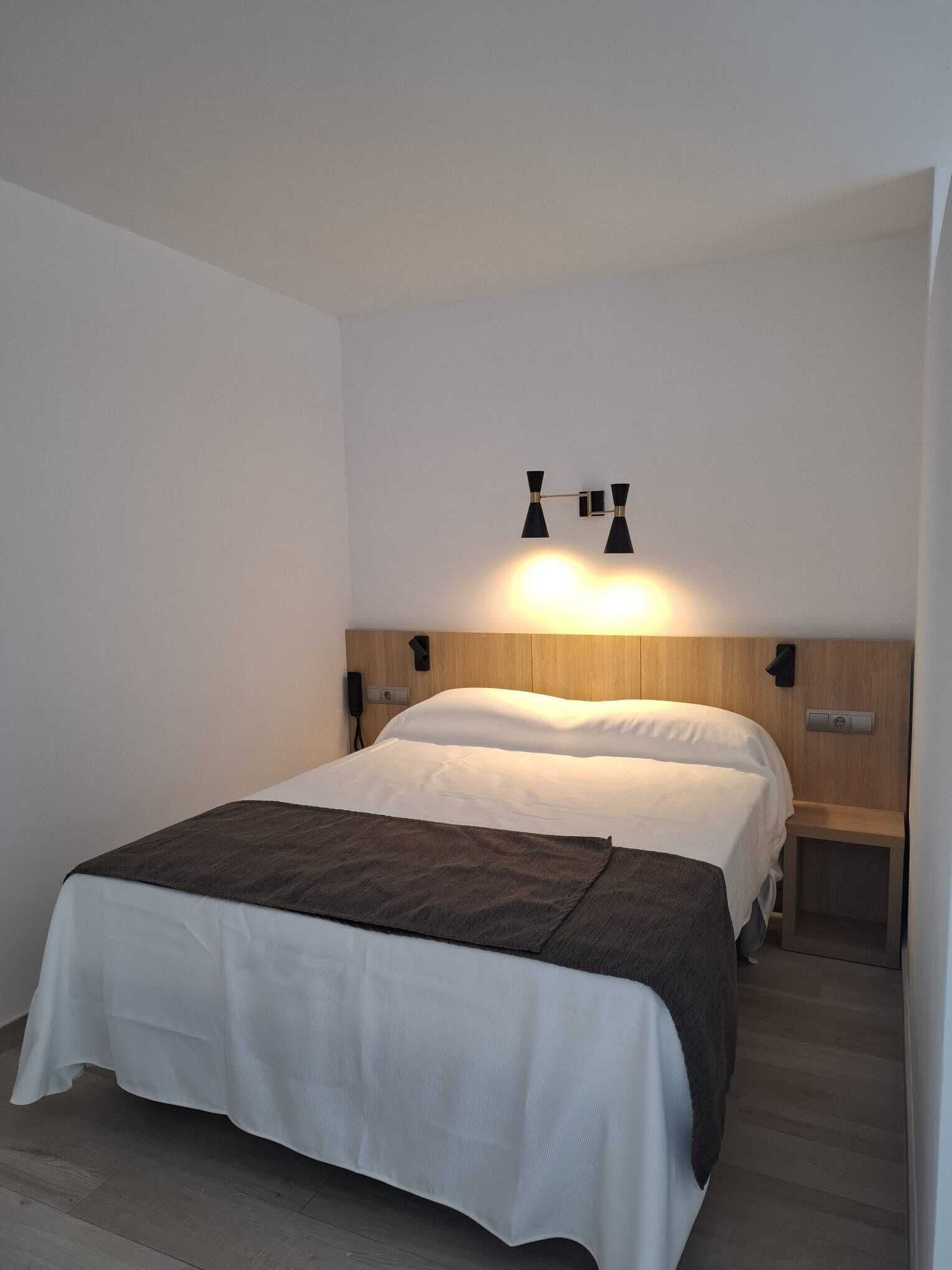 Una cama bien hecha con edredón blanco y manta marrón oscuro, un cabecero de madera y dos mesitas de noche fijadas a la pared con lámparas negras.