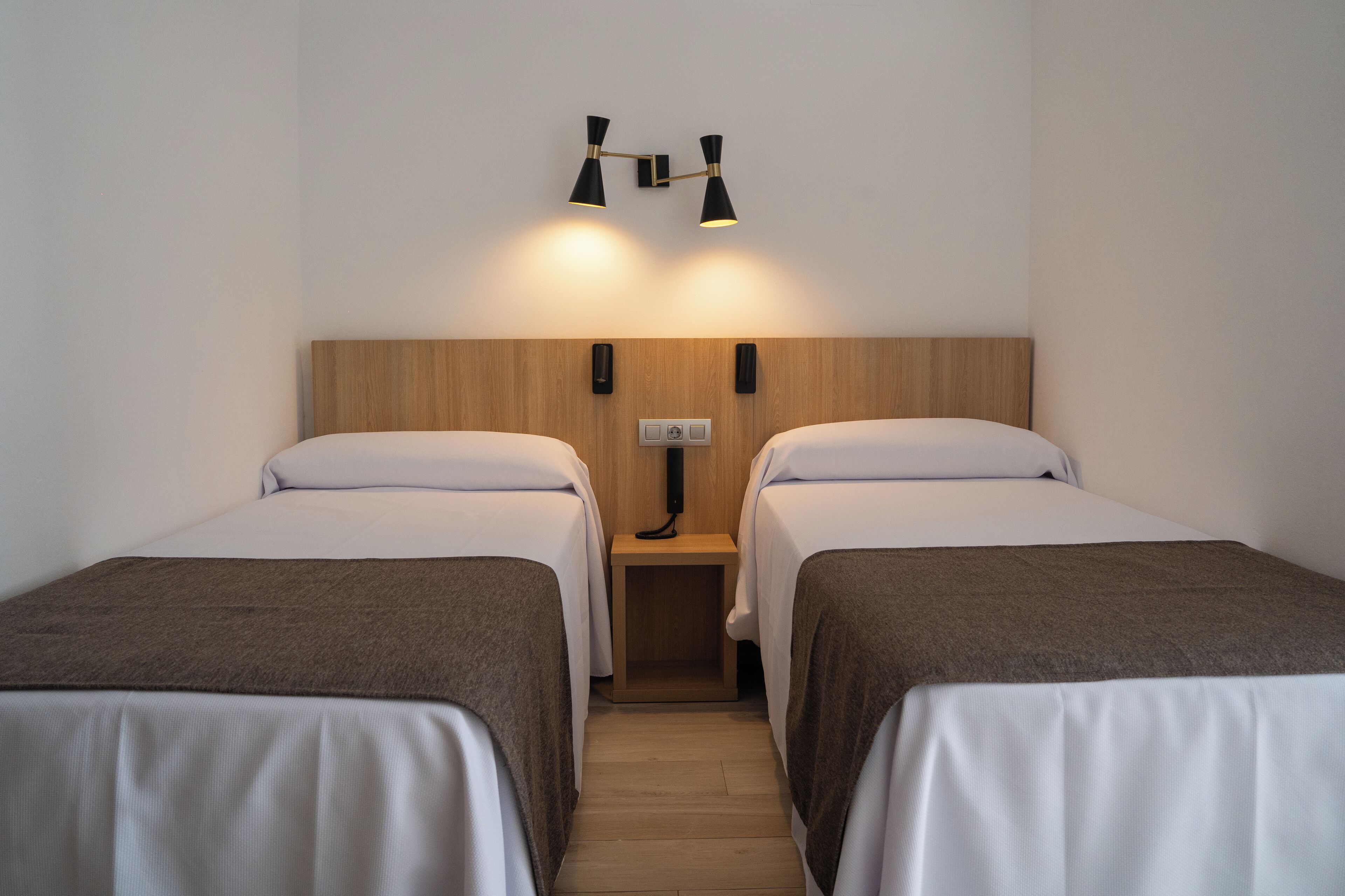 A hotel room with two single beds, a wooden headboard, a nightstand with a lamp, and a wall-mounted light fixture.