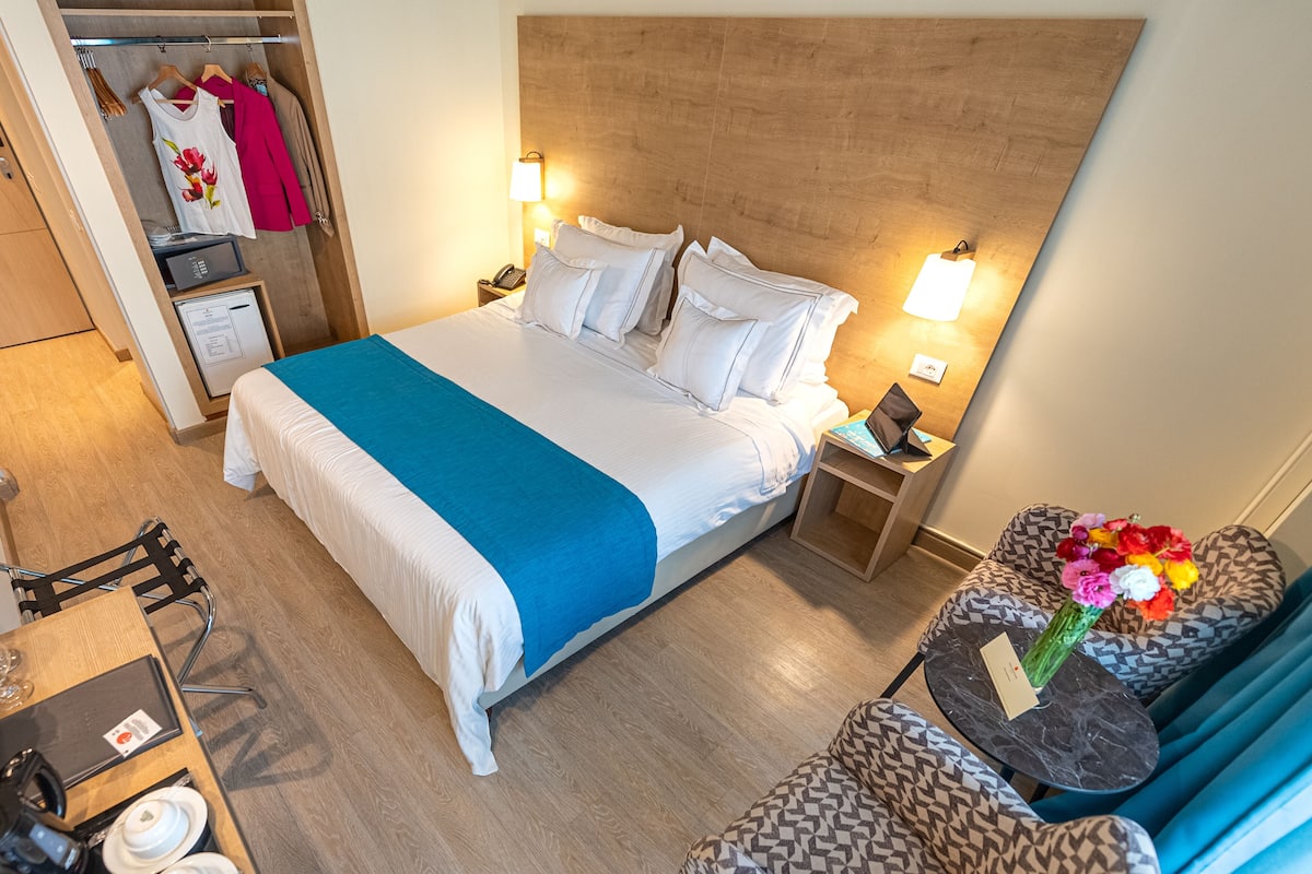 A hotel room with a large bed, bedside tables, a desk with a chair, a wardrobe, and a small seating area with a vase of flowers.