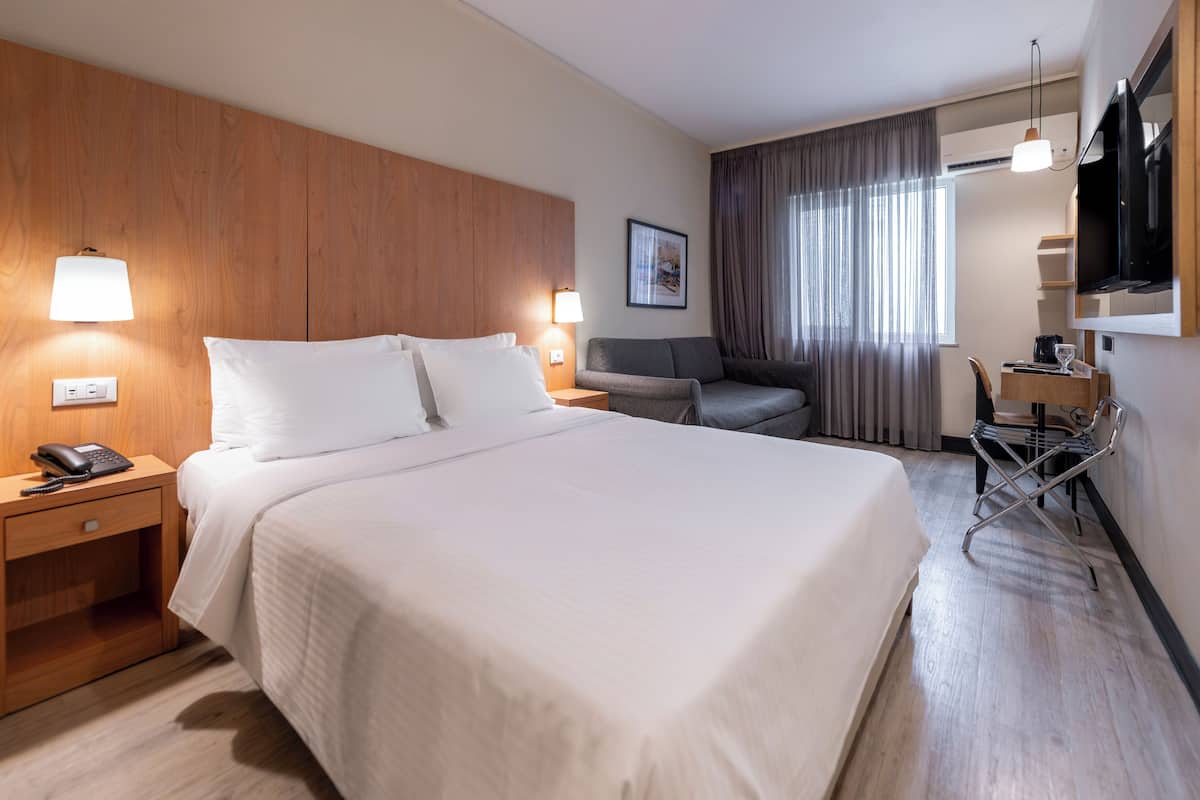 A hotel room with a large bed, bedside lamps, a desk with a chair, a TV, and a window with curtains.