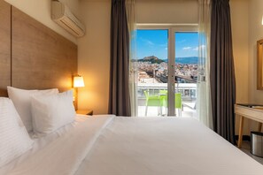 A hotel room with a bed, a desk, a lamp, and a view of a cityscape through a large window. - Polis Grand Hotel (Athens)