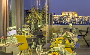 A restaurant with tables set for dining, potted plants, and a view of a historical building in the background. - Polis Grand Hotel (Athens)