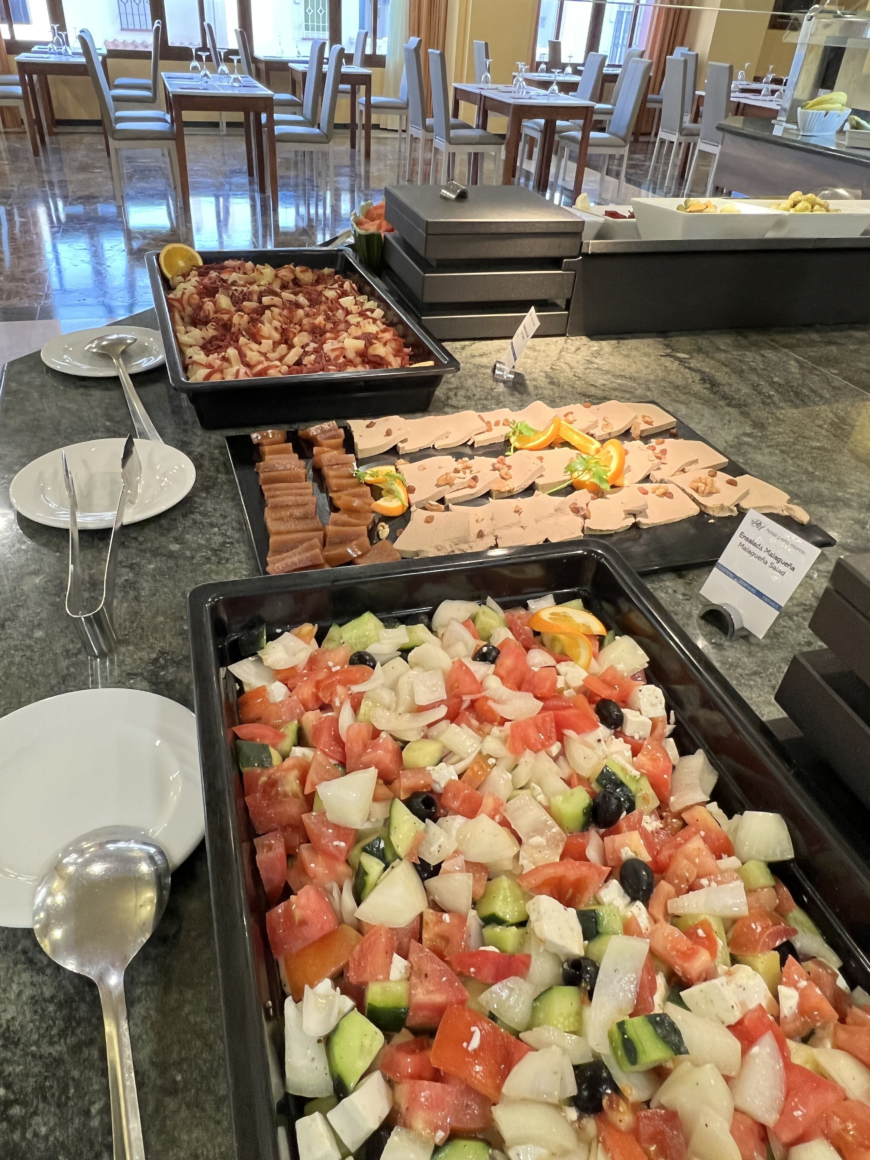 Un bufé con diversas opciones de comida, incluyendo una bandeja de verduras mixtas, un plato de ensalada de macarrones y una bandeja de frutas y nueces bañadas en chocolate.