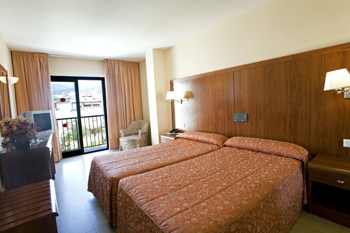 Een hotelkamer met een groot bed, een bureau met een computer, een stoel en uitzicht op het landschap door een schuifdeur.