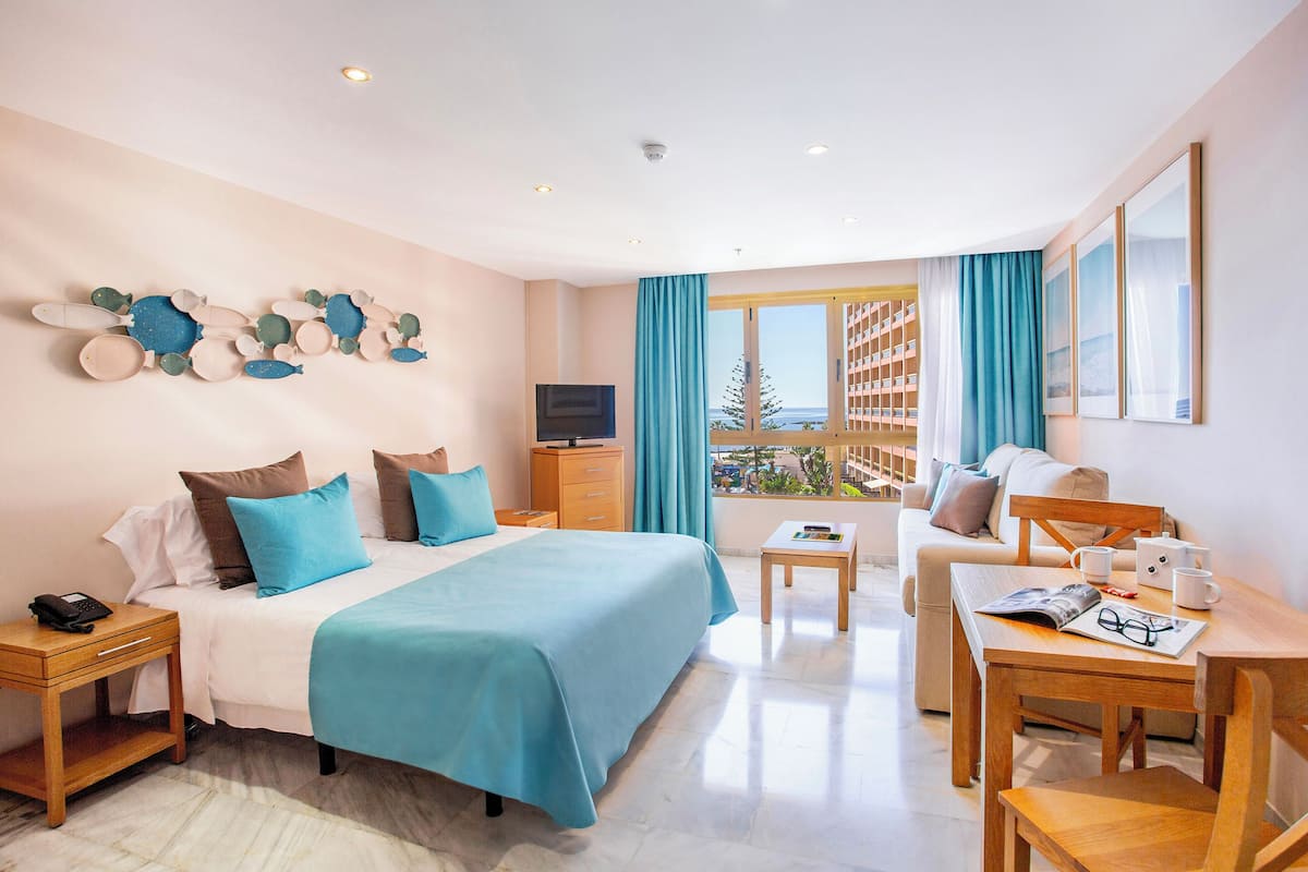 A hotel room with a bed, a TV, a desk with a chair, a window with curtains, and a balcony with a view.