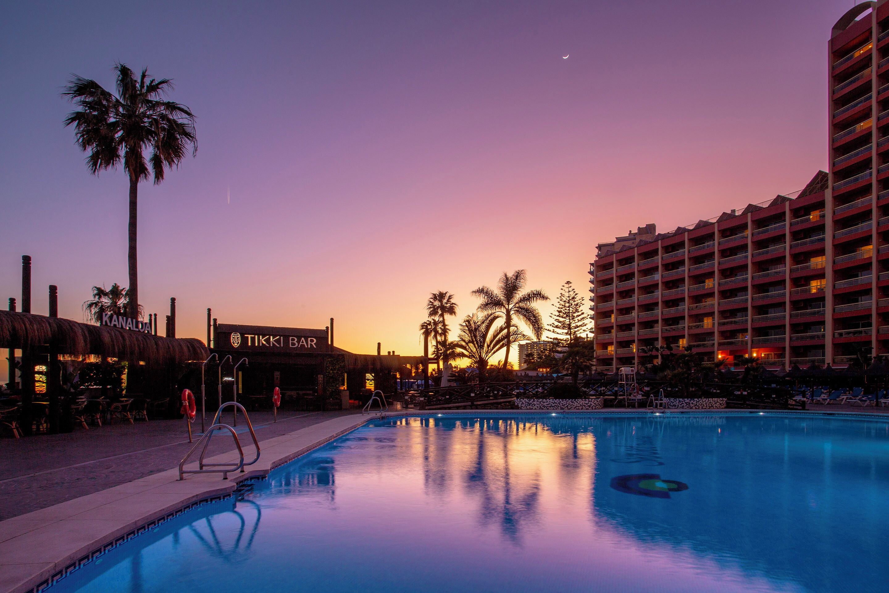 Een hotelzwembad met uitzicht op een zonsondergang, palmbomen en een bar genaamd 'TIKI BAR'.