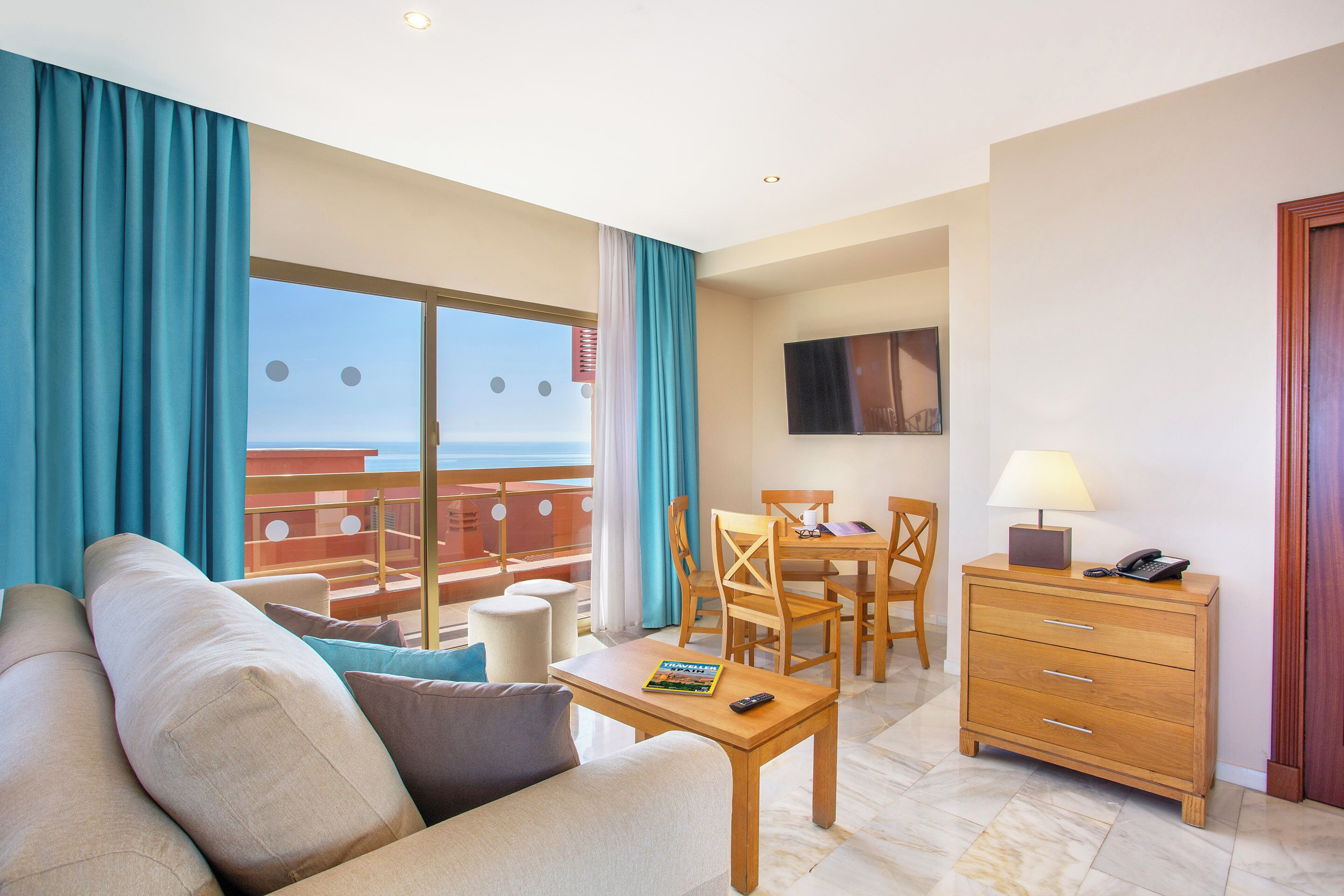 A hotel room with a balcony, a sofa, a dining table with chairs, a wooden dresser, and a TV mounted on the wall.