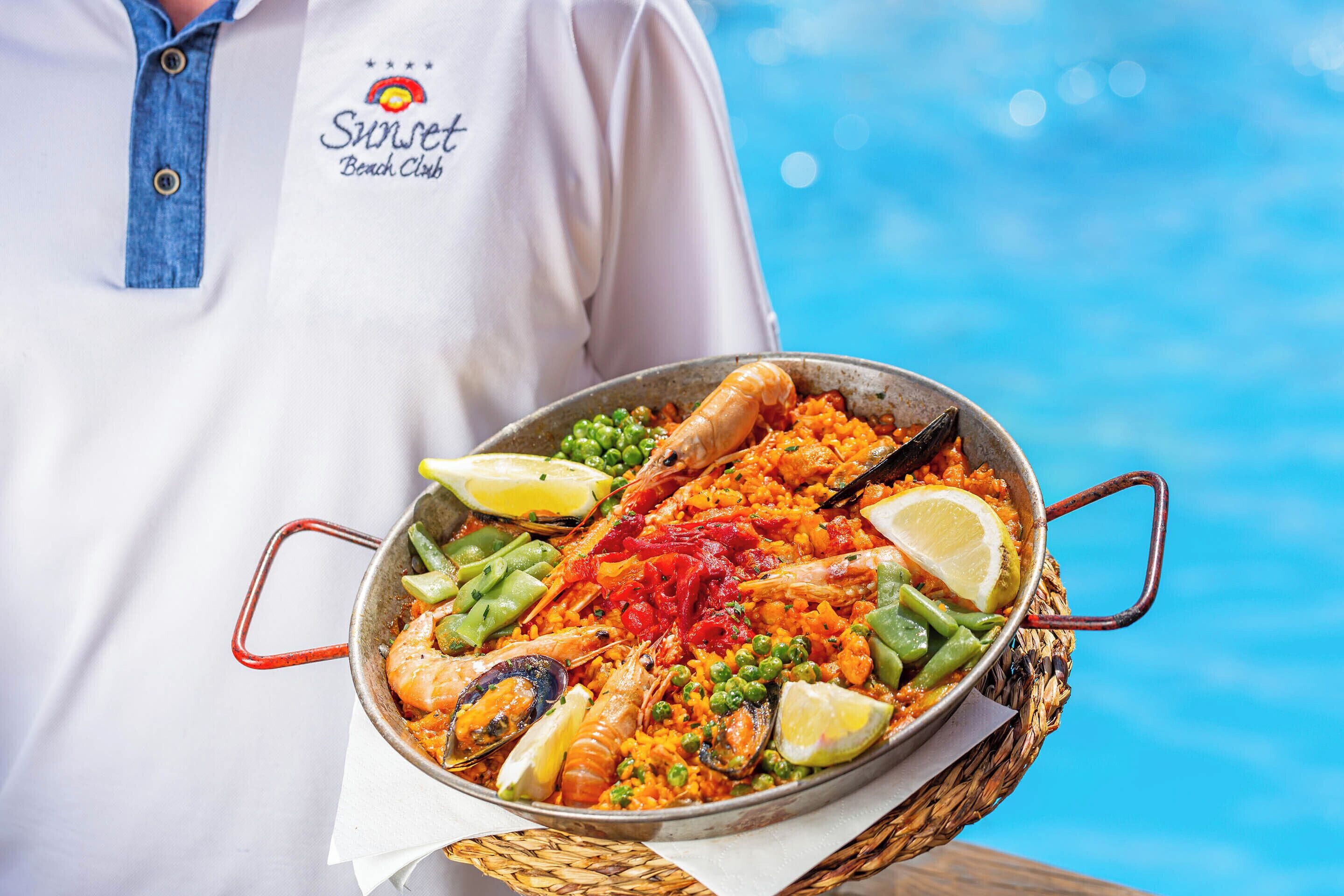 A person holding a large seafood paella with lemon slices and peas, standing by a pool.