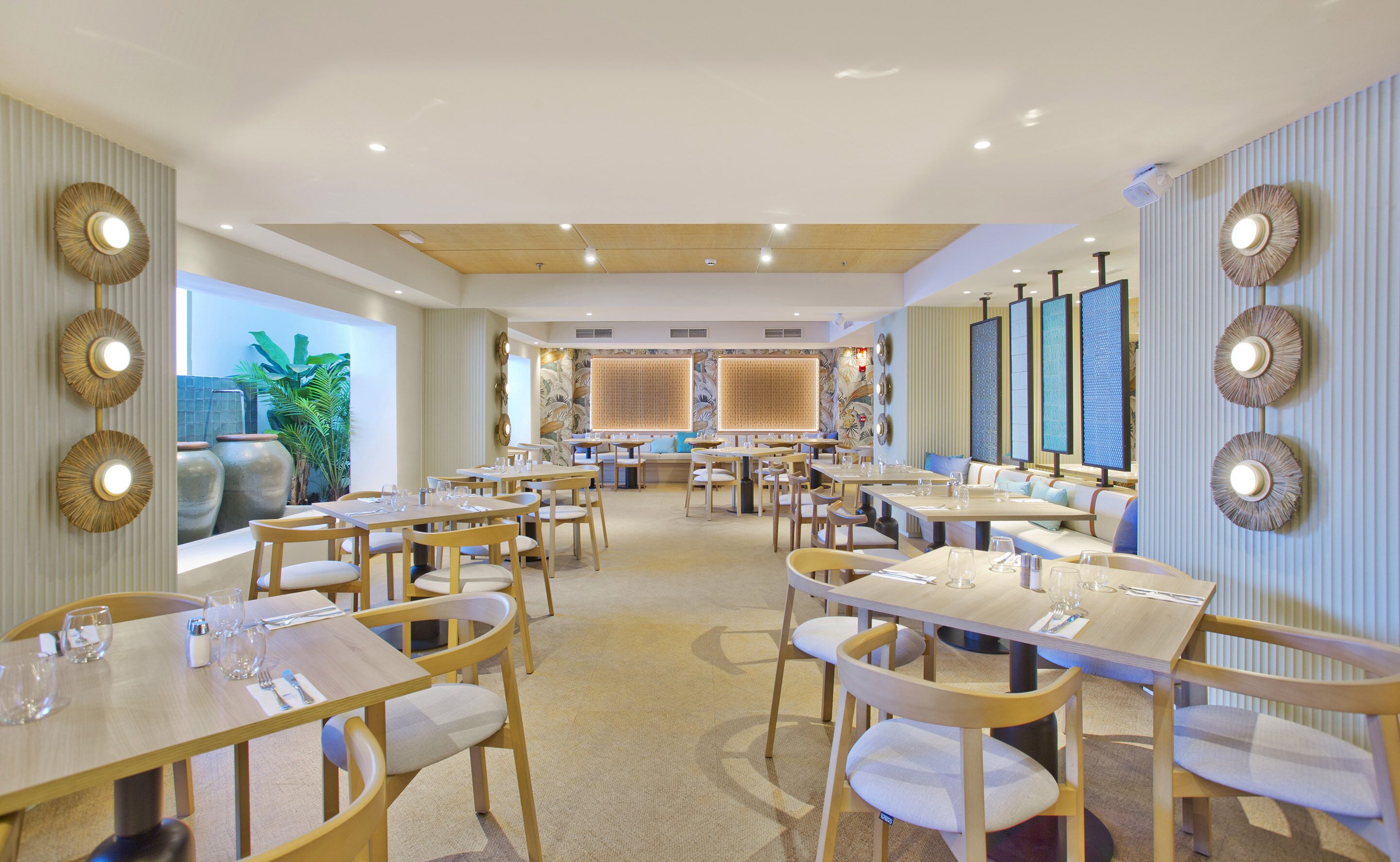 A spacious restaurant with multiple dining tables and chairs, a large plant in the background, and decorative wall panels.