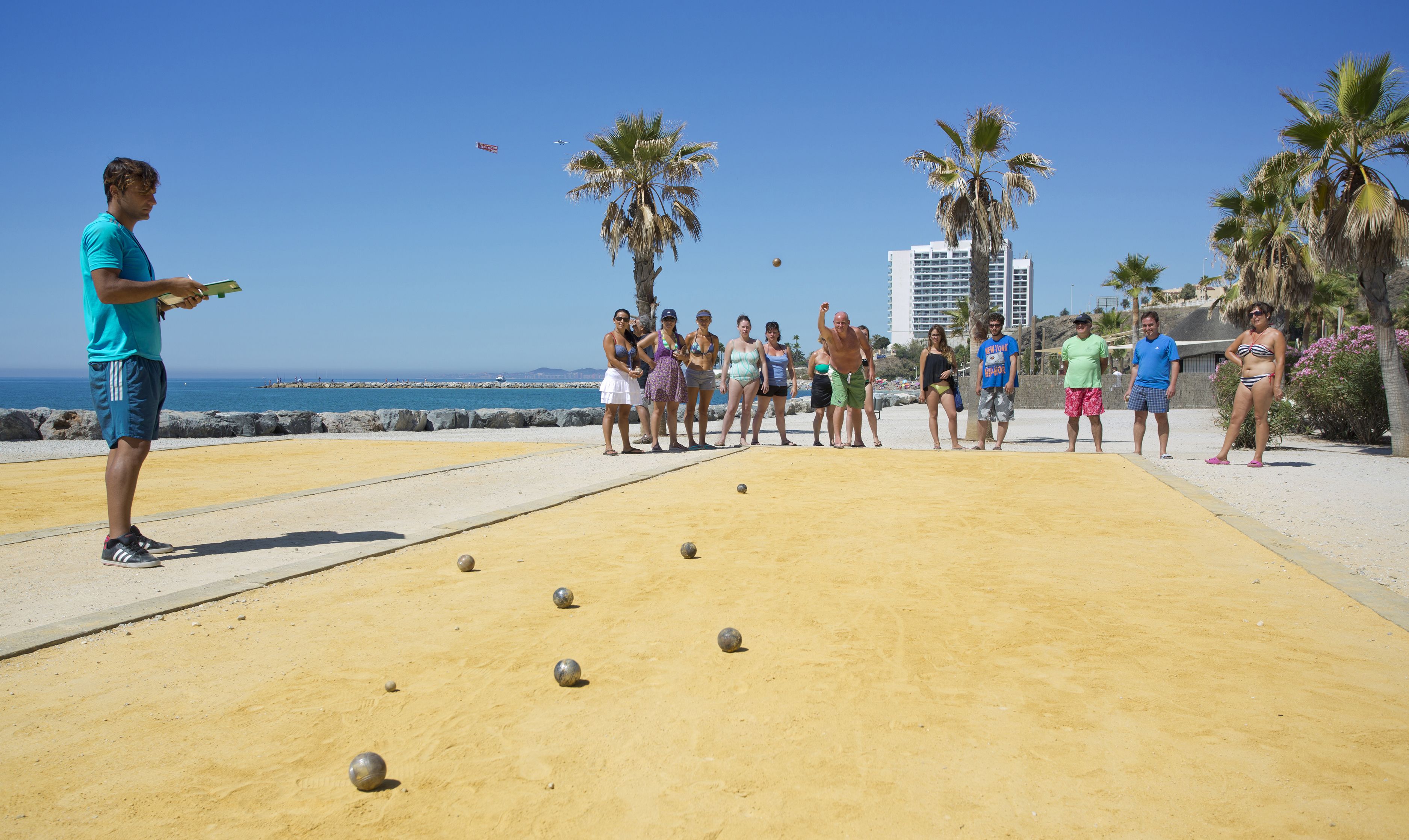 Um grupo de pessoas jogando bocha em quadra de areia, com uma praia e palmeiras ao fundo.
