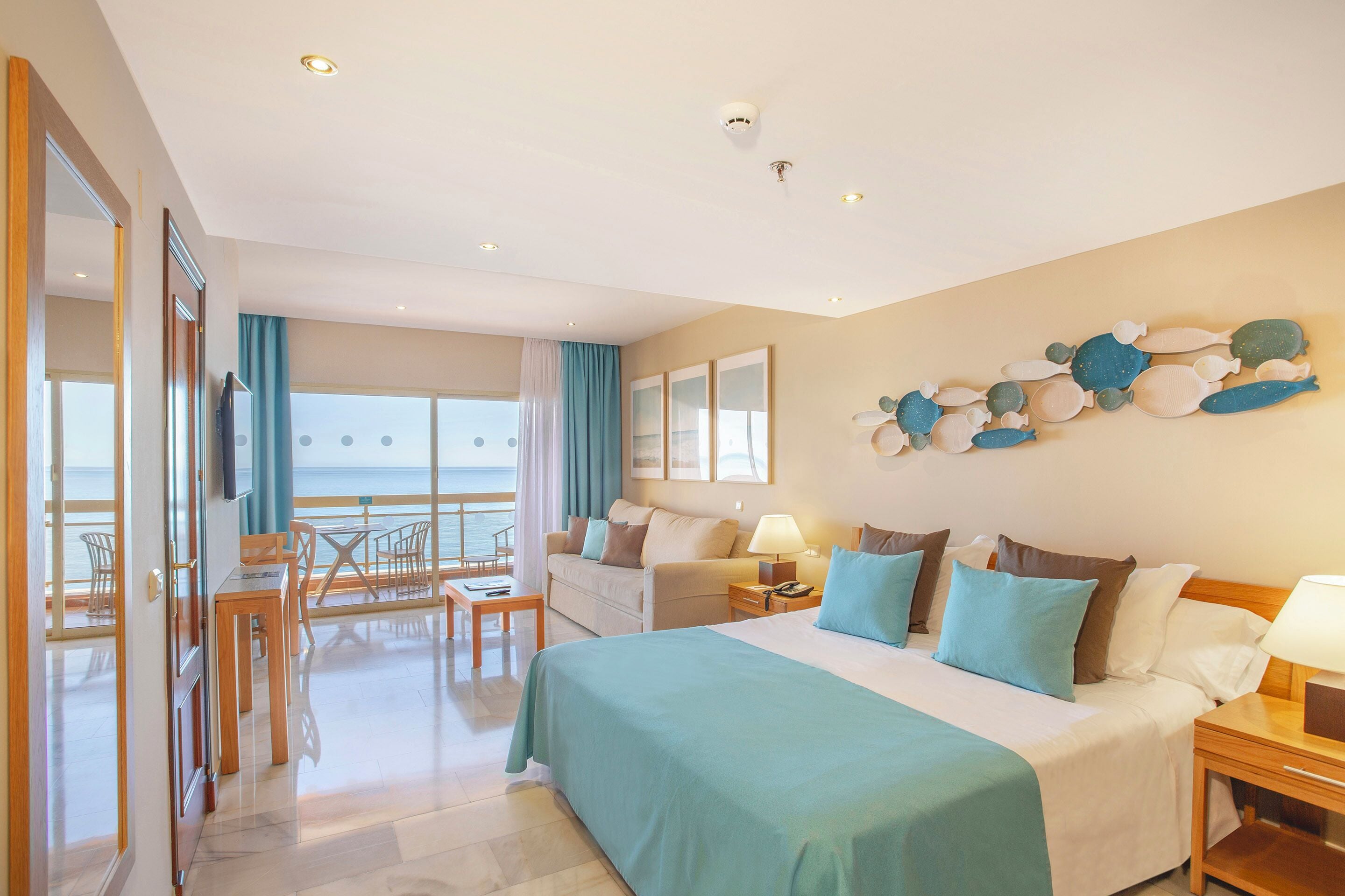 Habitación de hotel con cama, escritorio, silla, mesa, sofá, televisor y vista al mar.