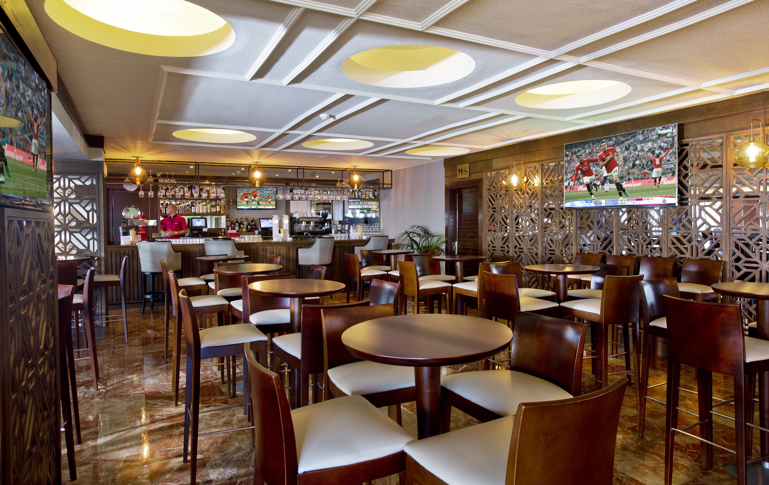 Un restaurante con mesas y sillas redondas, una zona de bar y una pared con temática deportiva.