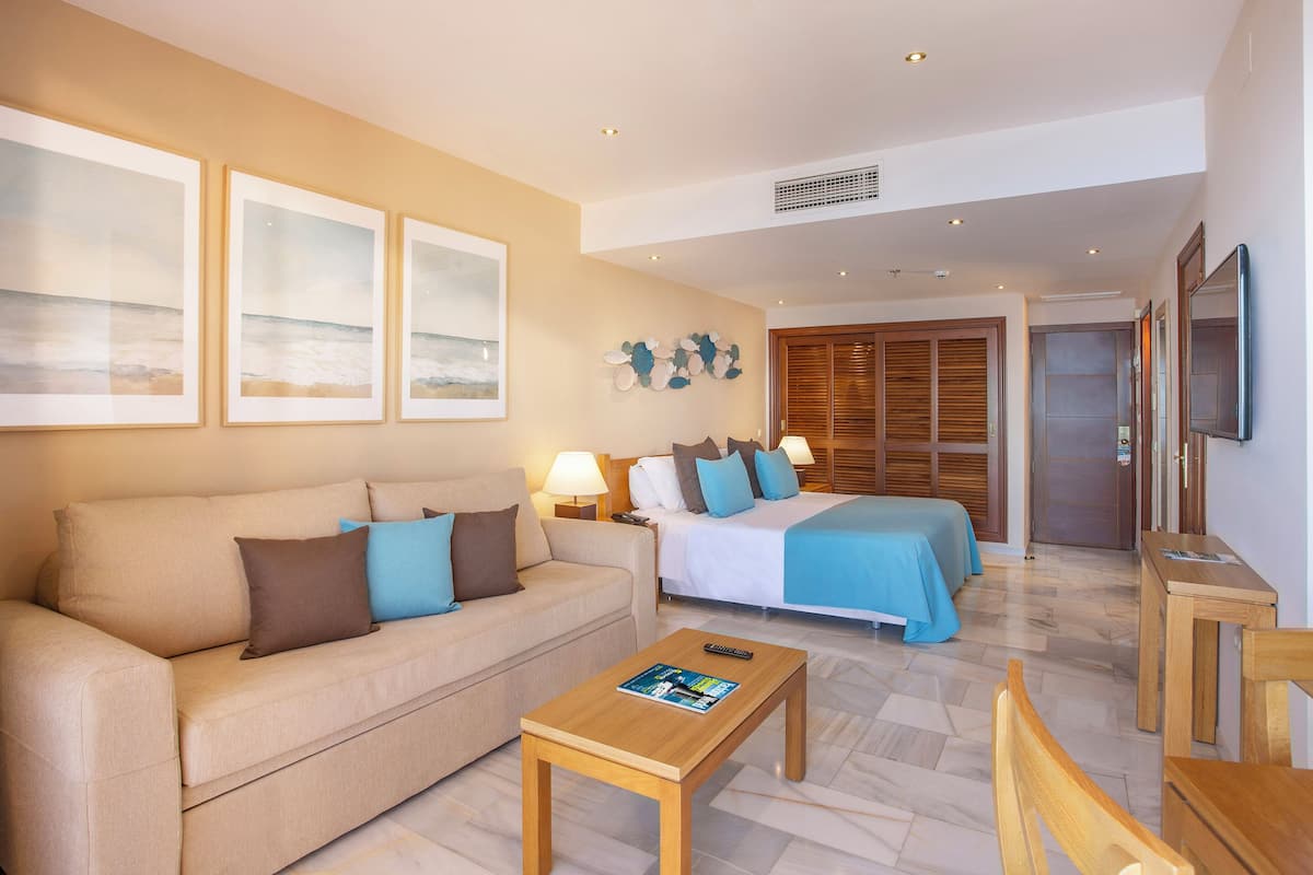 A hotel room with a sofa, a bed with a blue bedspread, a wooden coffee table, and a TV on the wall.