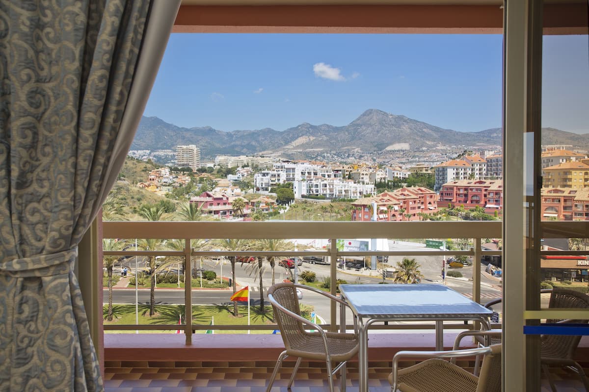 A balcony with wicker chairs and a table, overlooking a cityscape with buildings and mountains.