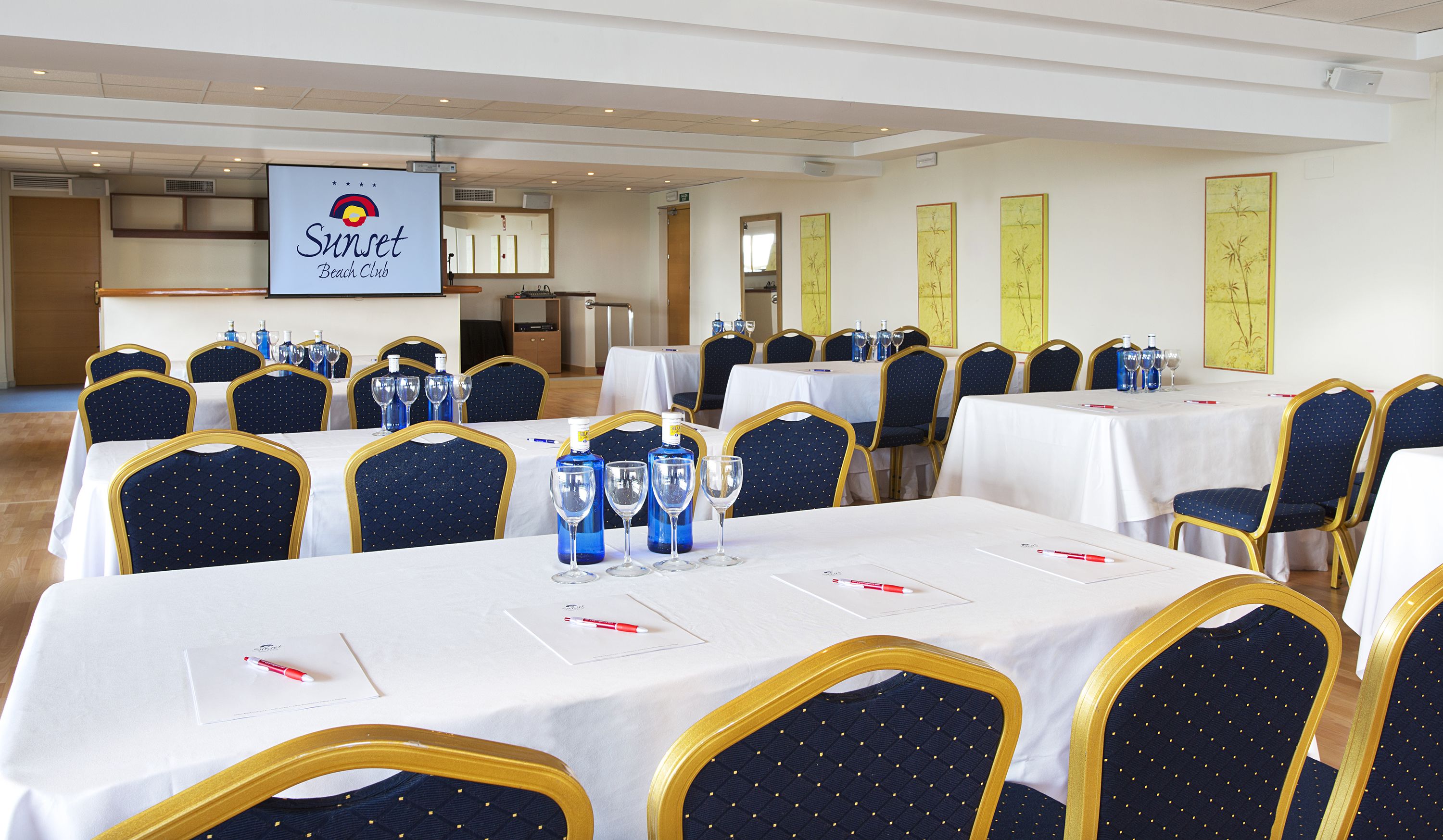 Un salón de conferencias con mesas dispuestas para una reunión, equipado con sillas y una pantalla para presentaciones.