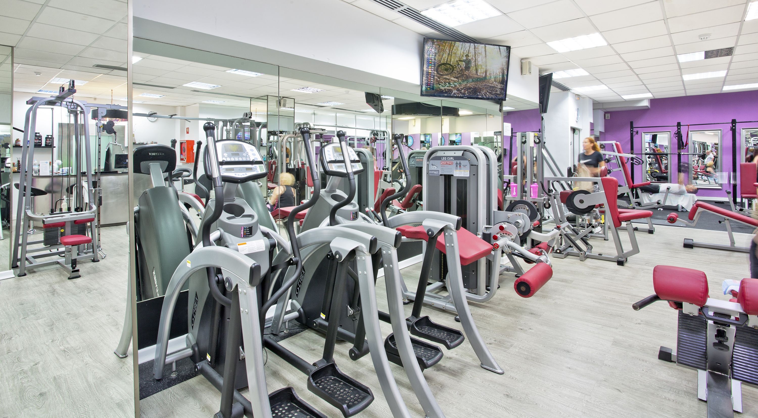 Un gimnasio con diferentes máquinas de ejercicio, espejos y una pantalla de televisión.
