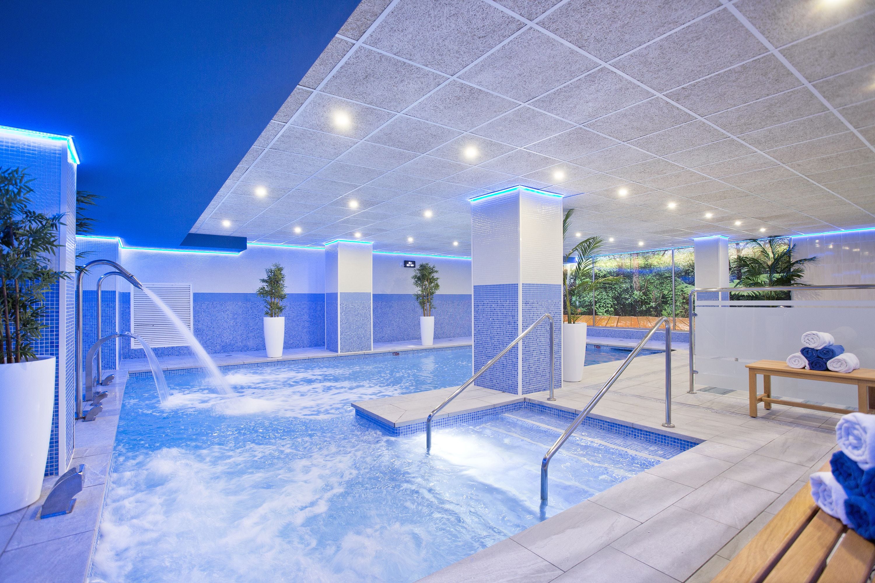 A modern indoor pool area with a hot tub, seating, and a view of the outdoors.