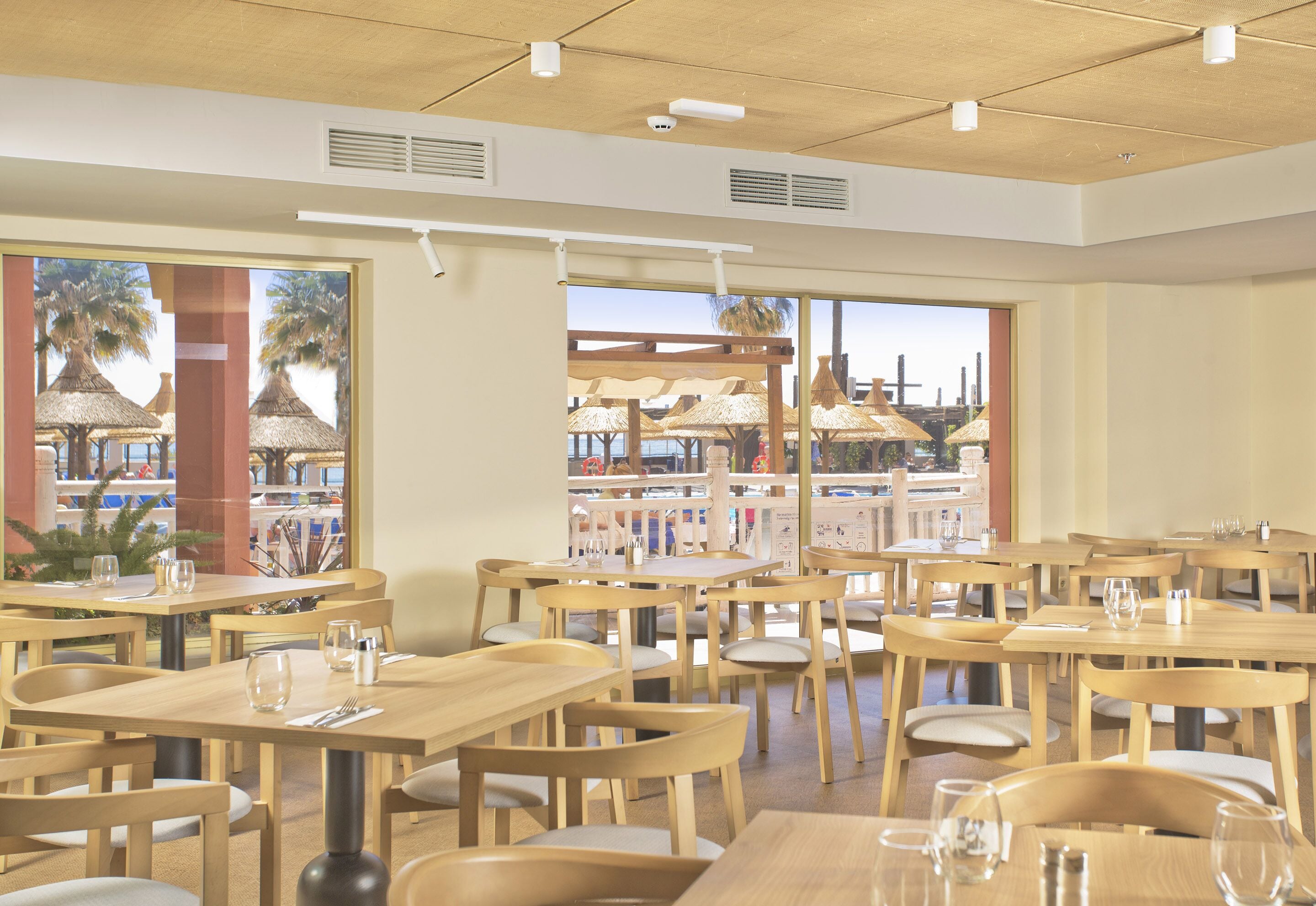 Un restaurant avec des tables et des chaises en bois, de grandes fenêtres et une vue sur les palmiers et les parasols à l’extérieur.