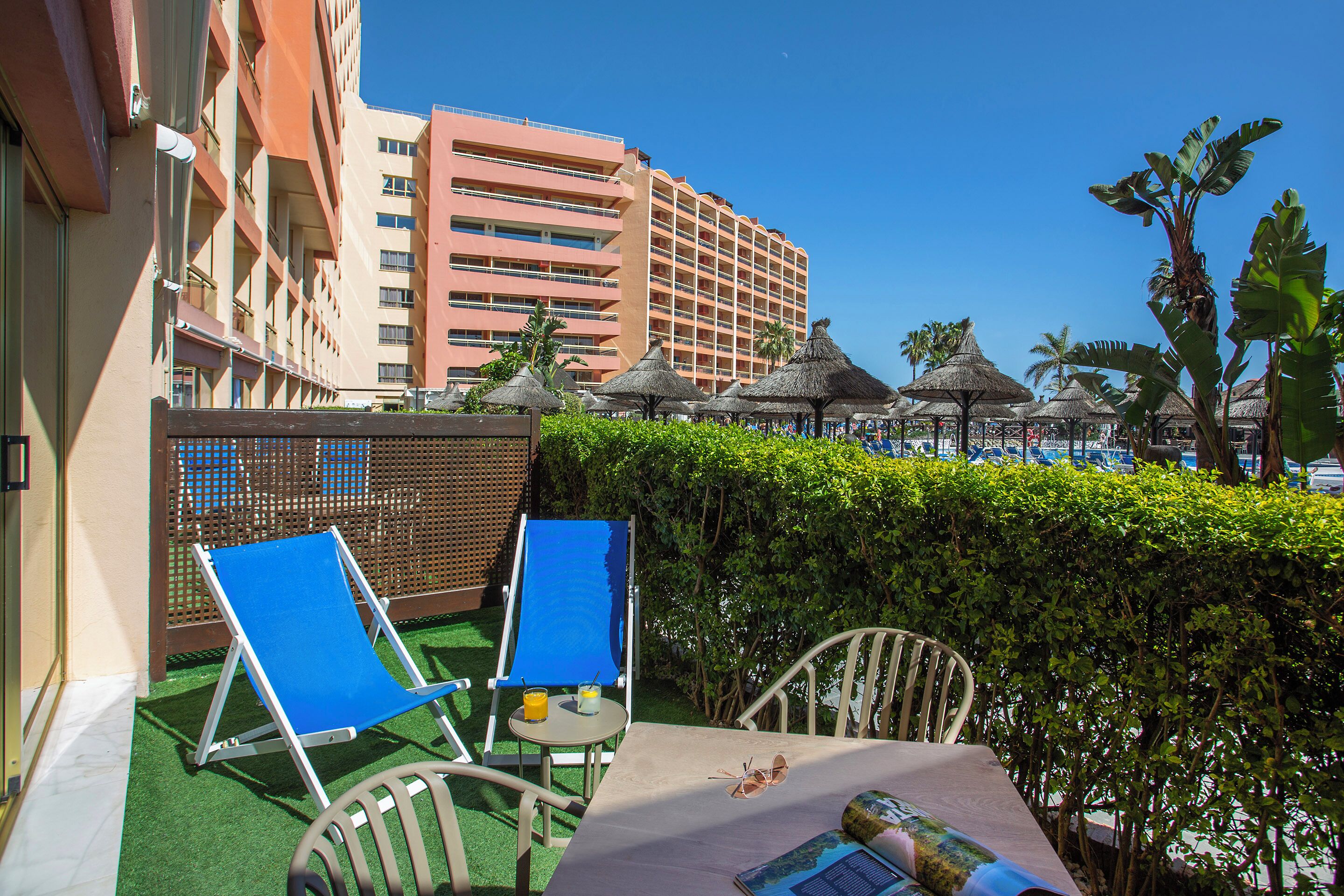 A hotel balcony with two blue lounge chairs, a table with a drink and sunglasses, and a view of a tall building in the background.