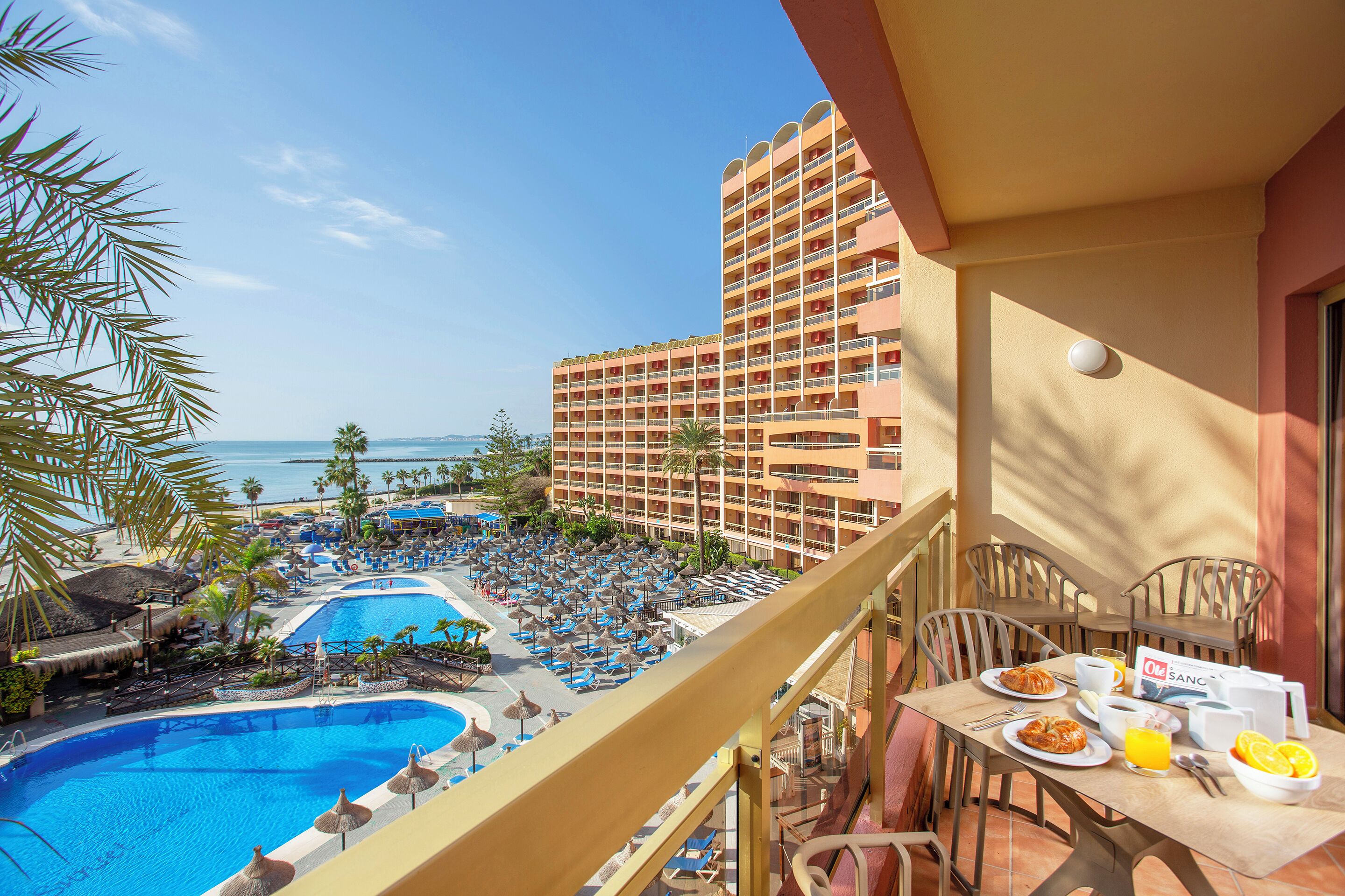 Un balcon d’hôtel avec vue sur la piscine, une table à manger où le petit-déjeuner est servi, et une vue sur l’océan et les bâtiments de l’hôtel.
