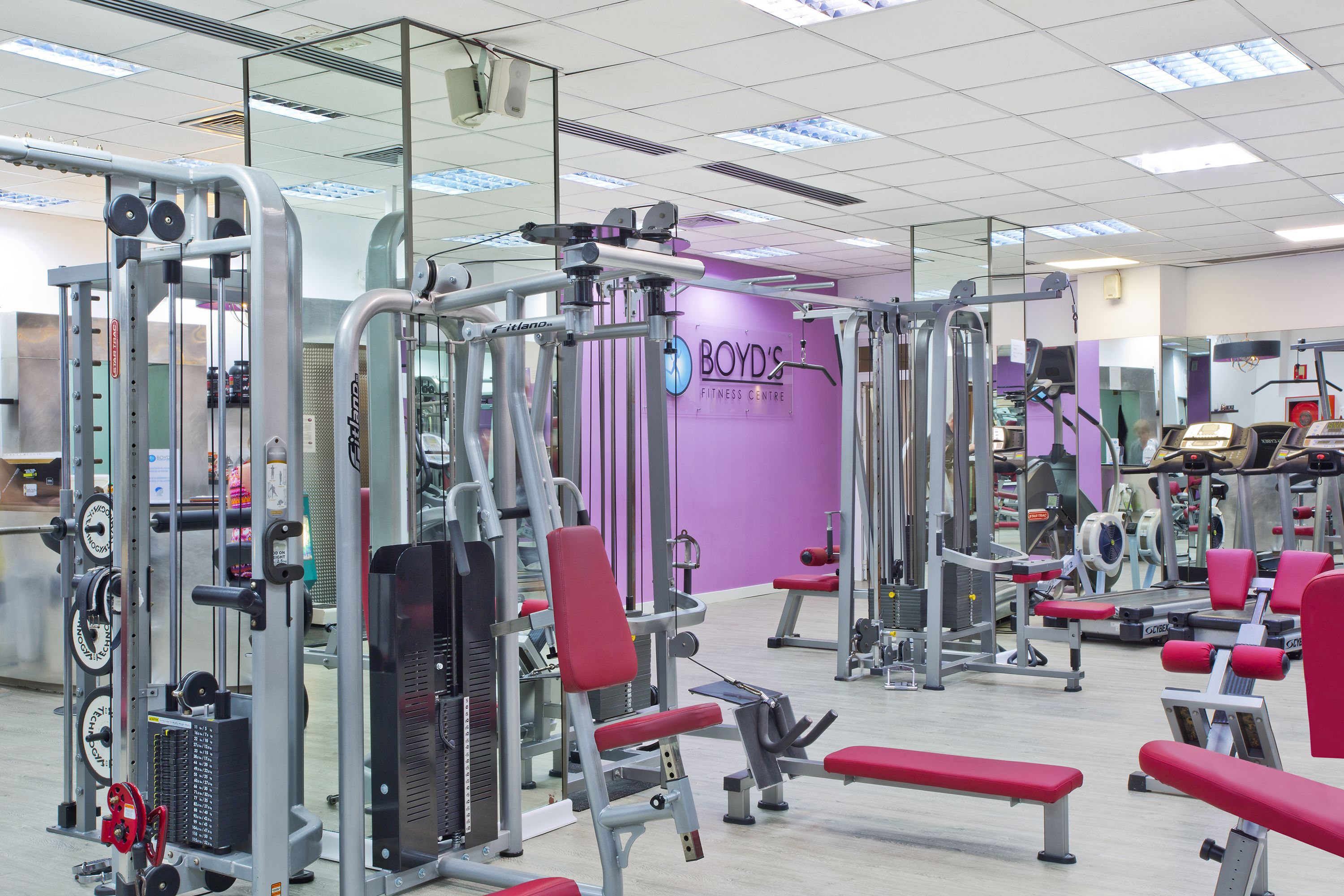 Un gimnasio con diversas máquinas de ejercicio, bancos y una pared morada con el nombre "BOYD'S FITNESS CENTRE".