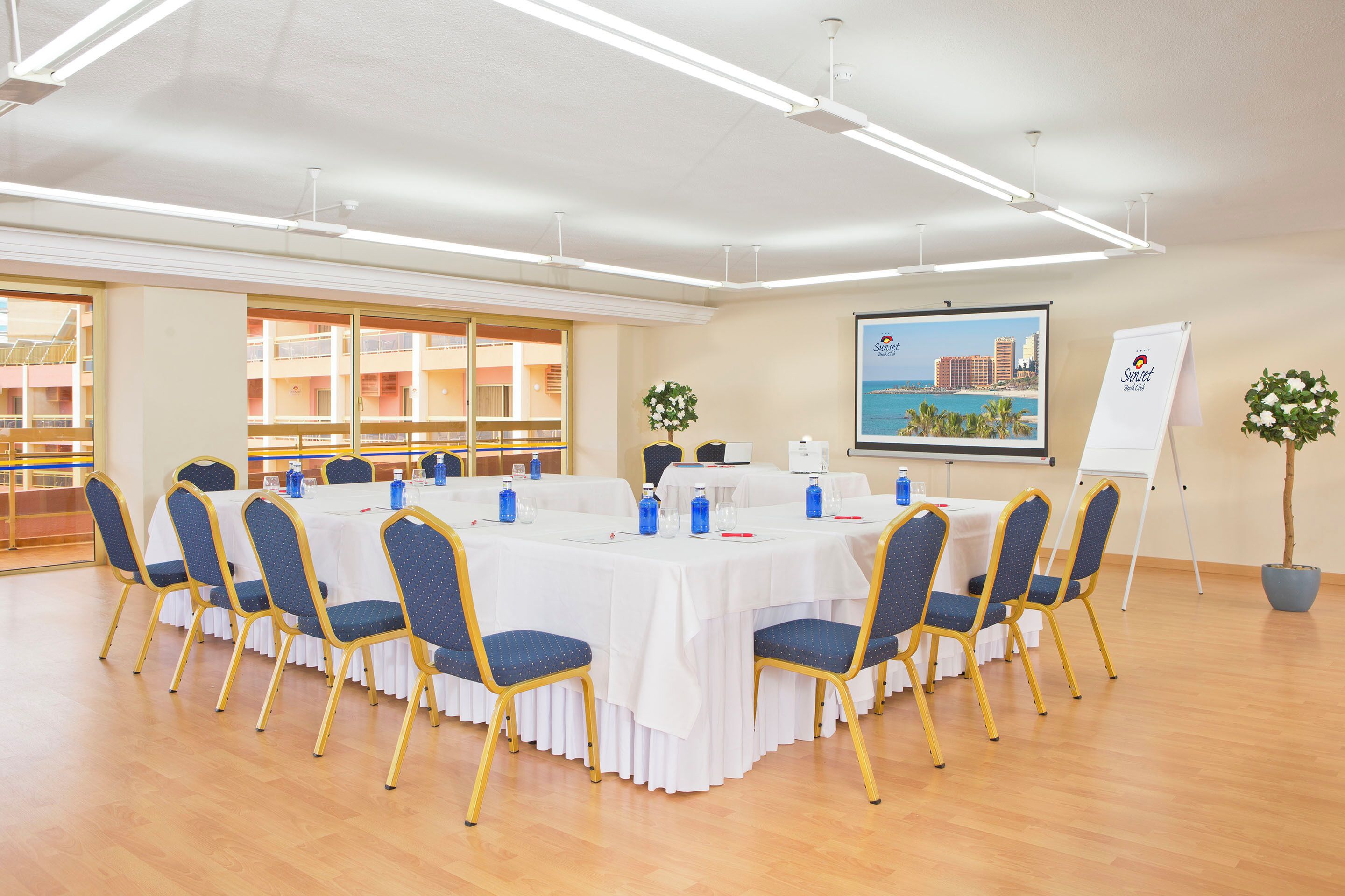 Un salón de conferencias con una mesa redonda, sillas azules, manteles blancos y un cartel con un paisaje urbano en la pared.