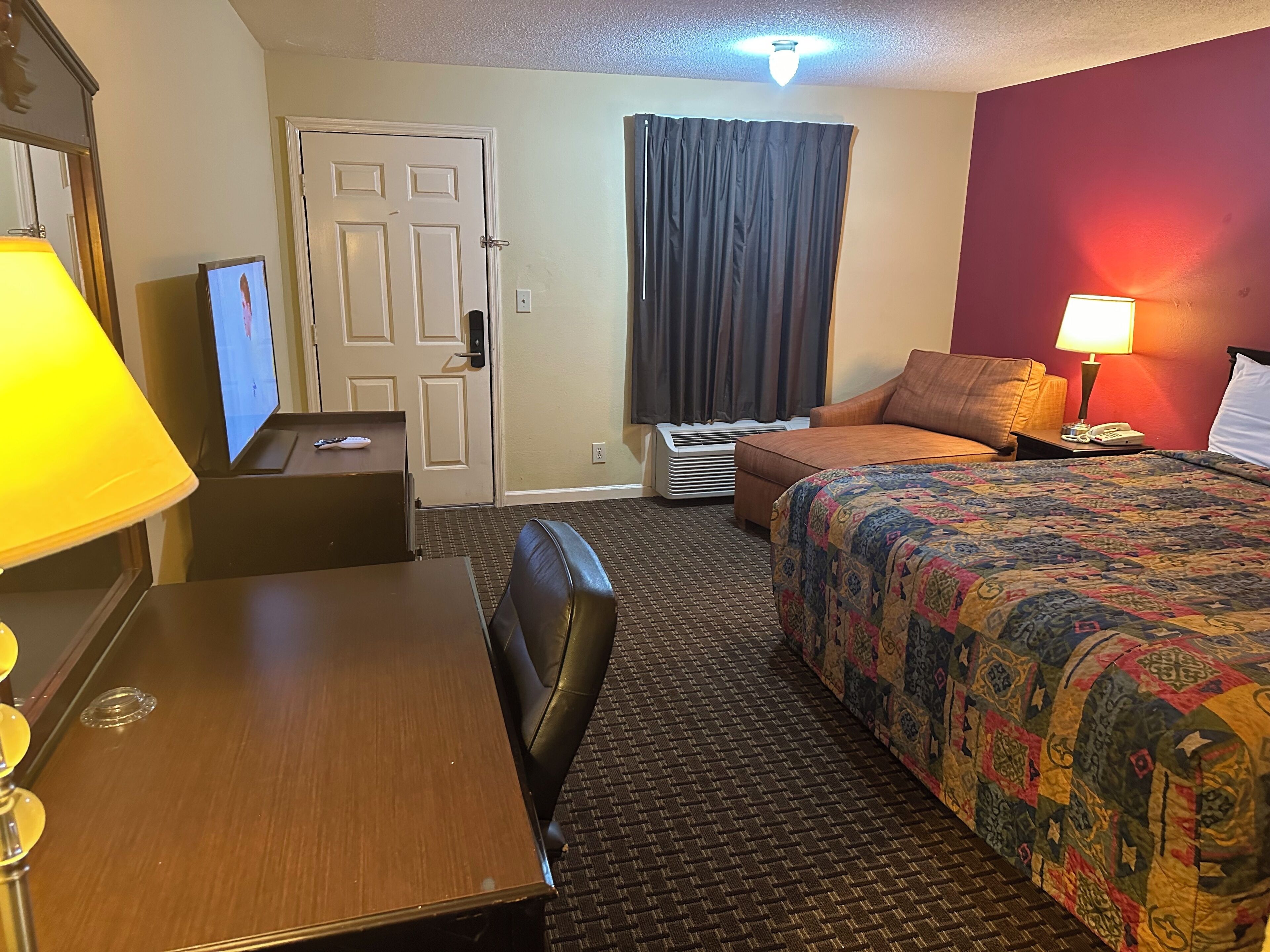 Quarto de hotel com uma cama, uma escrivaninha, uma cadeira, uma televisão, um abajur e uma janela com cortinas.