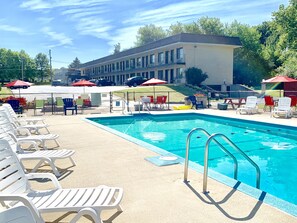 A swimming pool with lounge chairs and umbrellas, surrounded by a fence and a building in the background. - Geneva Wells Inn (Lake Geneva)