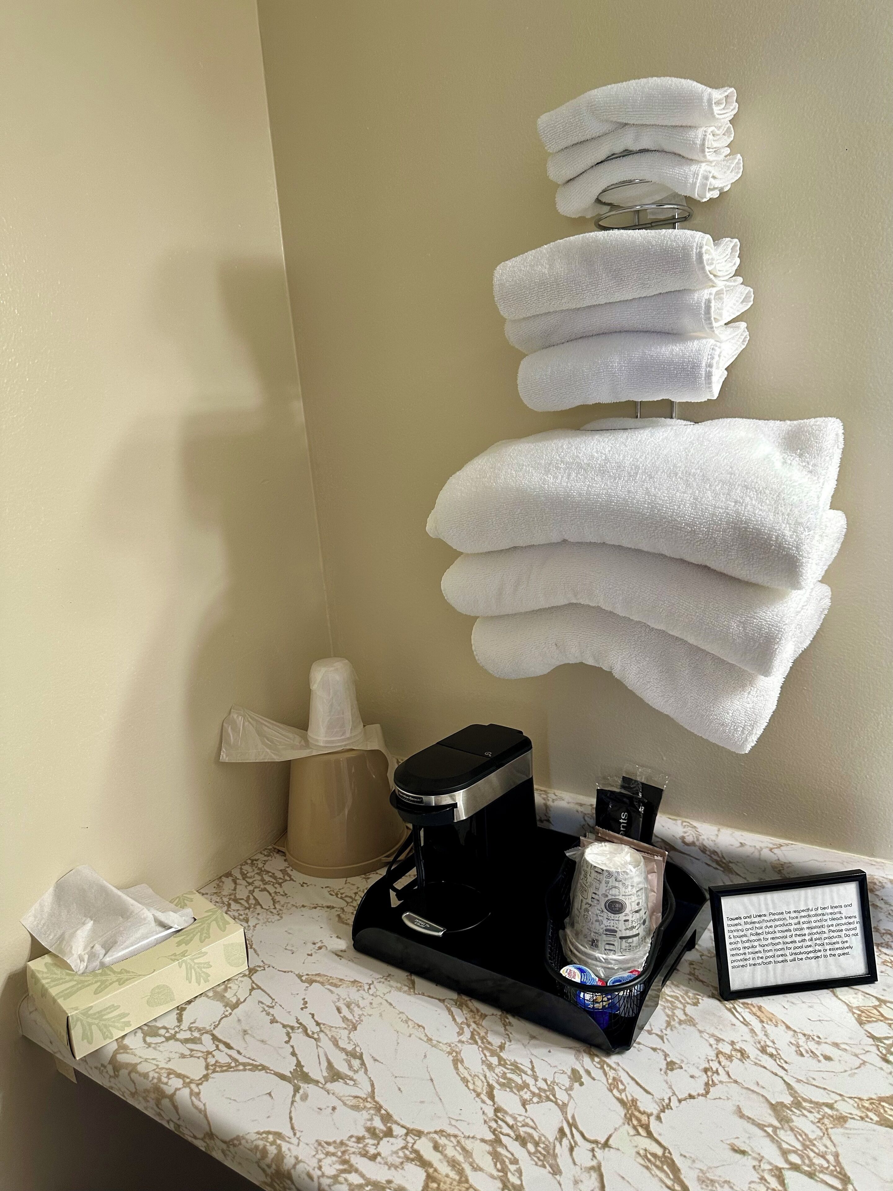 A bathroom counter with a coffee maker, a tissue box, and a stack of white towels.